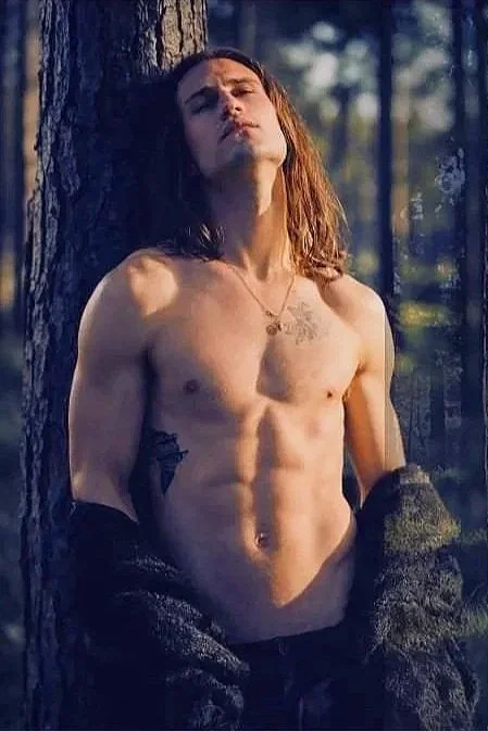 A shirtless man with long hair standing outdoors in a forest, with sunlight filtering through the trees, leaning against a tree trunk. Photography by Riaan Swiegelaar