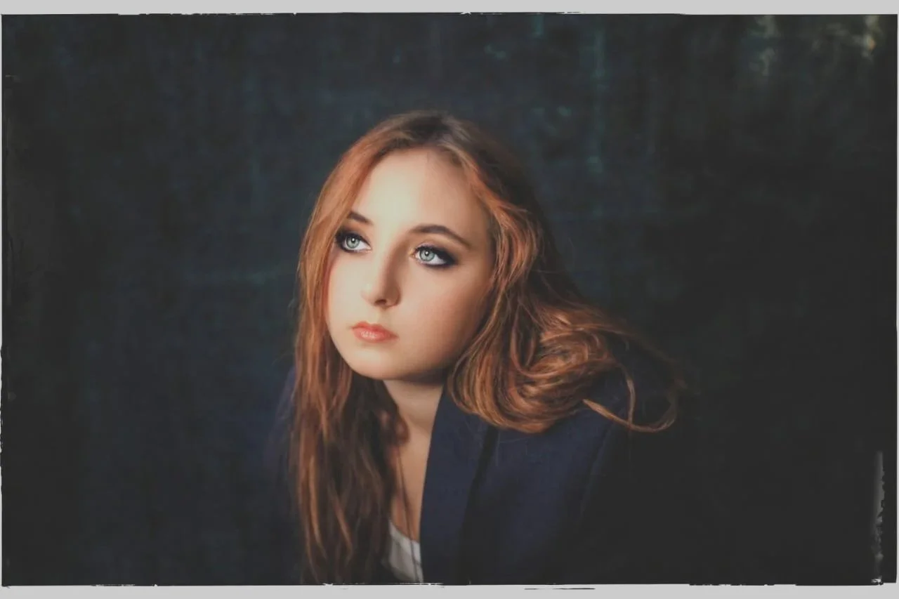 A young woman with long reddish-brown hair and blue eyes gazes upward and to the left in a dark setting. Photography by Riaan Swiegelaar