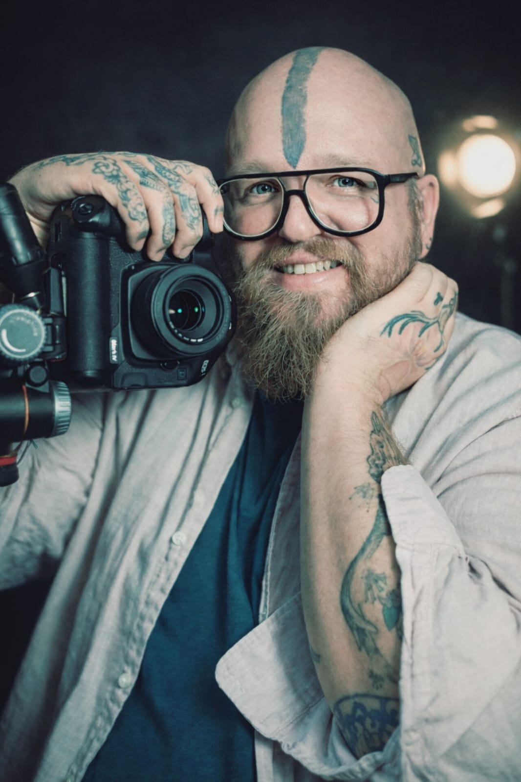 Bio pic of Riaan Swiegelaar . A smiling man with tattoos and glasses holding a camera, wearing a beige shirt and dark t-shirt, in a photography studio with dark background and studio light.