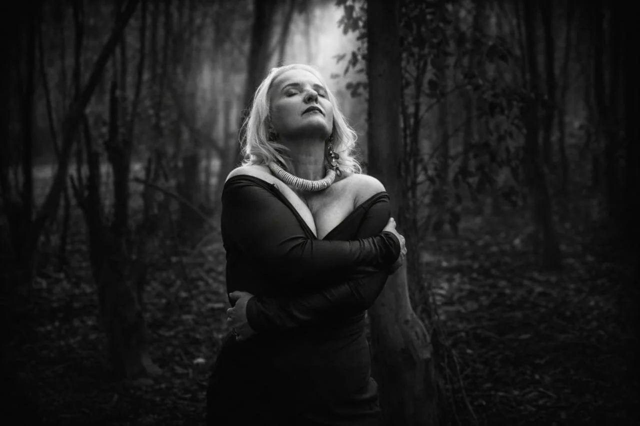 Black and white photo of a woman standing in a forest, wearing an off-shoulder dress and a statement necklace, with her eyes closed and arms crossed. Photography by Riaan Swiegelaar