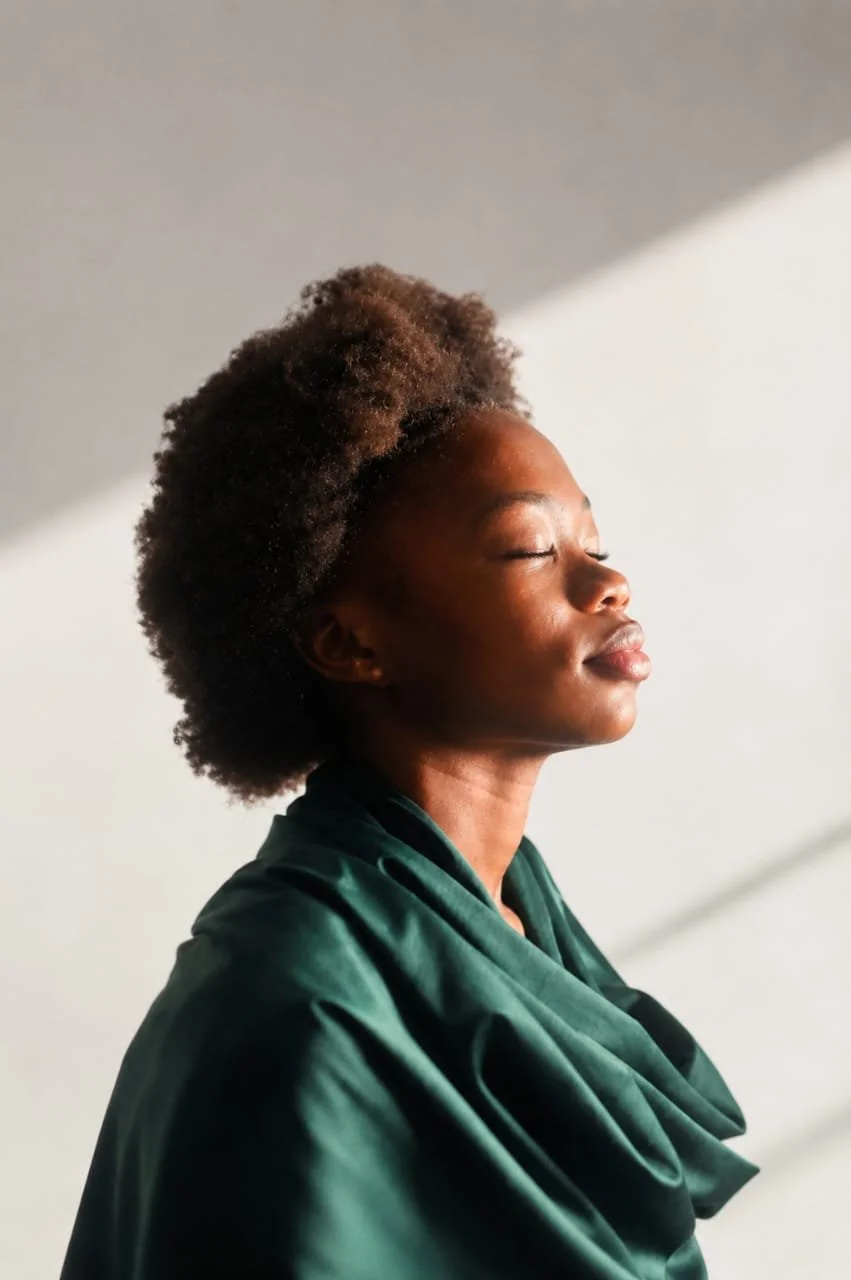 A woman with natural curly hair and a dark green satin robe, eyes closed, face lifted towards the sunlight, appearing peaceful and content. Photography by Riaan Swiegelaar
