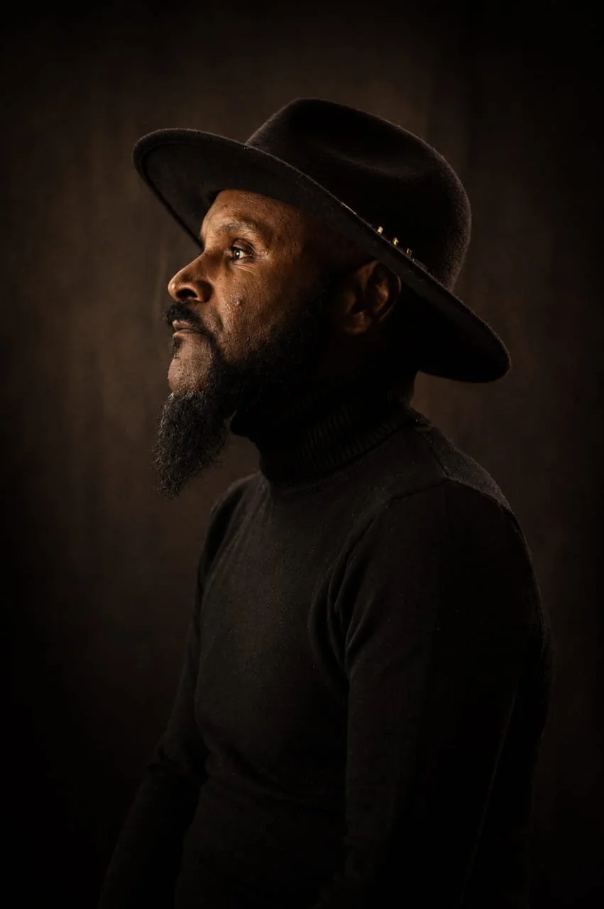 A man in profile wearing a black turtleneck and wide-brimmed black hat, looking to the left against a dark background.