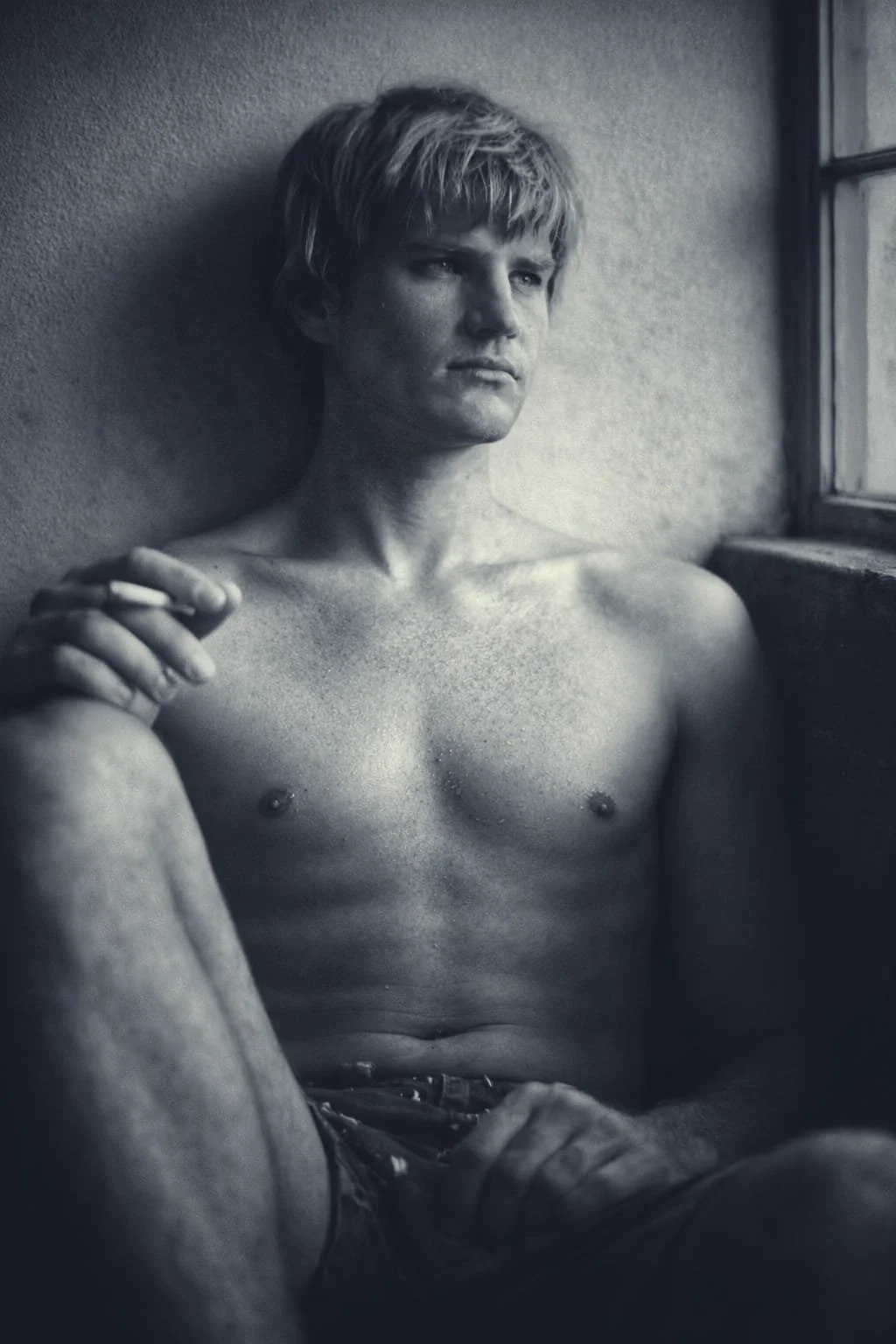 Black and white photo of a shirtless young man with short blonde hair, sitting against a wall near a window, holding a cigarette in his right hand, looking contemplative. Photography by Riaan Swiegelaar