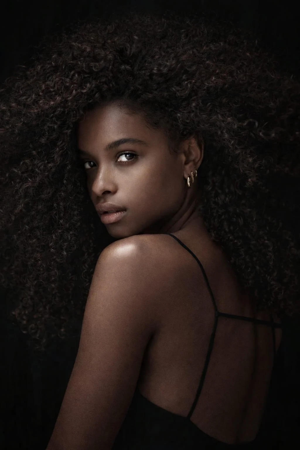 A woman with dark skin and voluminous curly hair looks over her shoulder, wearing a black spaghetti strap top and gold hoop earrings. Photography by Riaan Swiegelaar