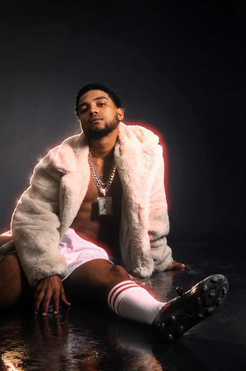 A young man sitting on the floor wearing a light-colored fur coat, pink shorts, white soccer socks with red stripes, black soccer cleats, and layered chain necklaces with pendants, against a dark background with a red glow outline.