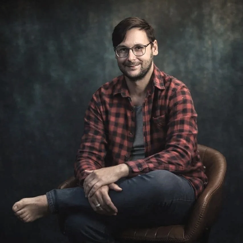 A man with glasses, dark hair, and a beard smiling, sitting barefoot on a brown leather chair, wearing a red and black plaid shirt over a gray T-shirt, against a dark textured background. Photograph by Riaan Swiegelaar