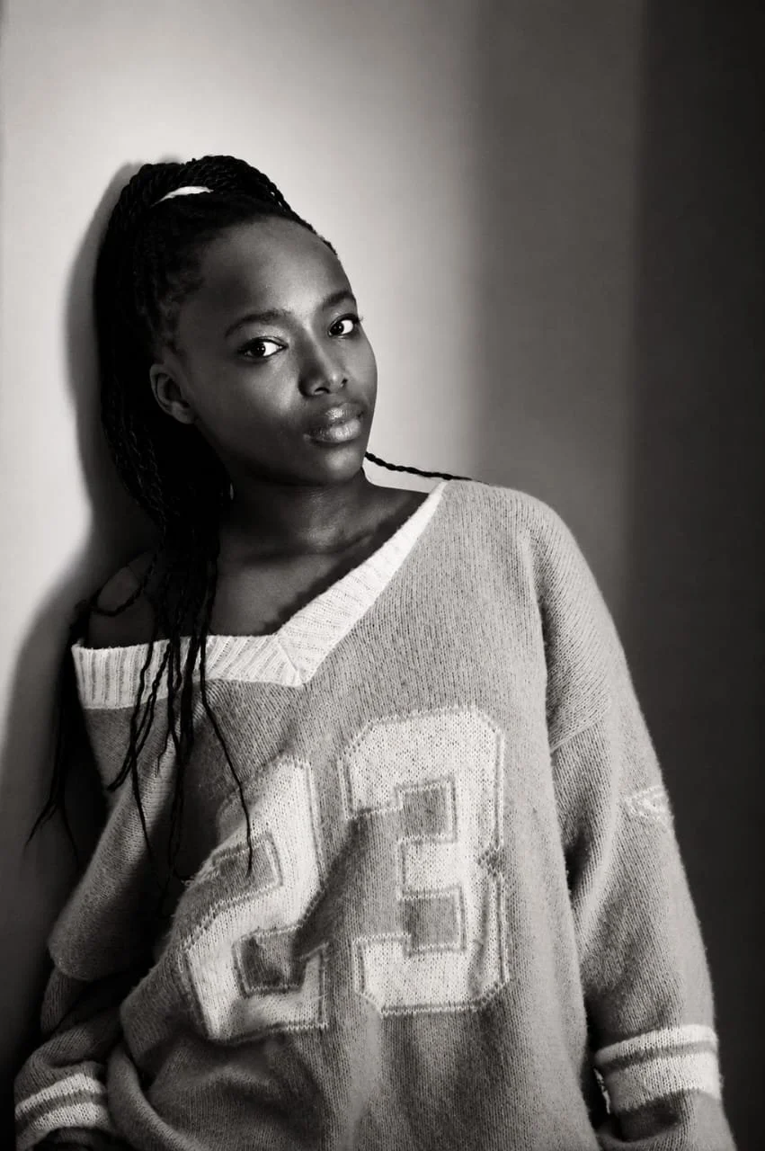 A young woman with braided hair leaning against a wall, wearing a Sports jersey with the number 23, looking at the camera in black and white. Photography by Riaan Swiegelaar