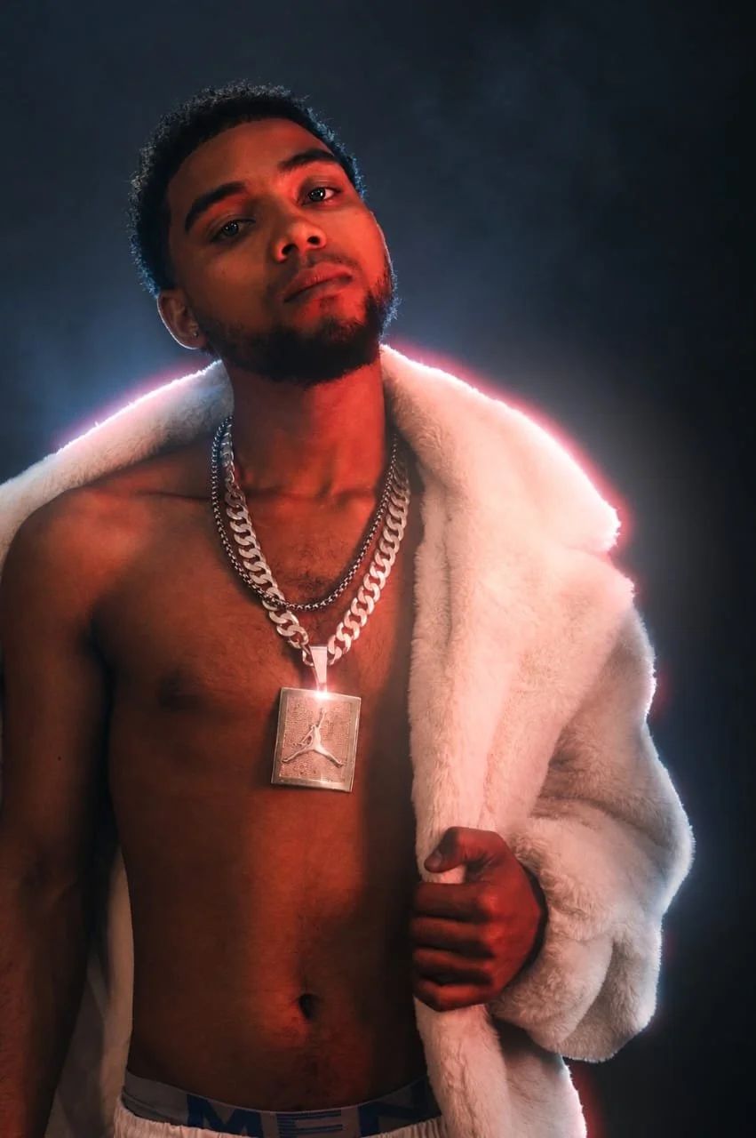 Photography by Riaan Swiegelaar. A shirtless young man wearing chain necklaces, a white fur coat draped over his shoulder, and boxer shorts, posed against a dark background with dramatic lighting.