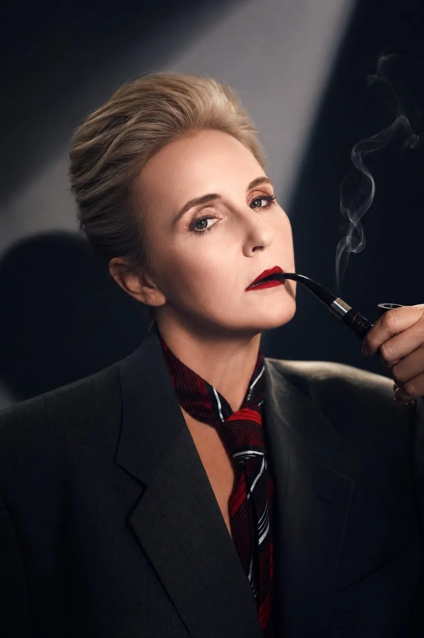 A woman with short blonde hair, wearing a dark blazer and a striped neck scarf, is smoking a pipe with smoke rising from it.