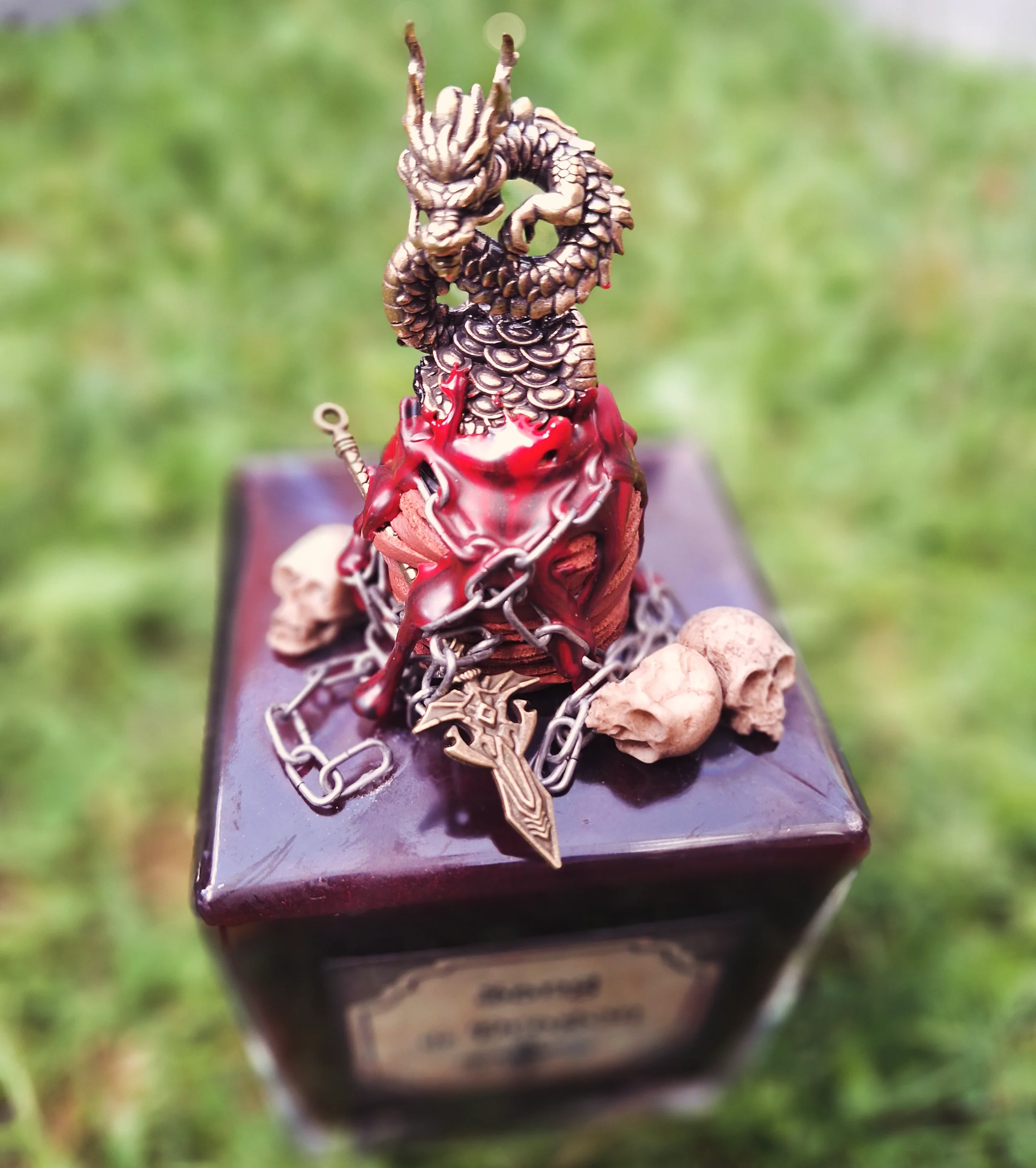 Une figurine de dragon en métal doré, entourée de chaînes, posée sur un coffret en bois foncé, avec trois roches autour, dans un environnement en plein air verdoyant.