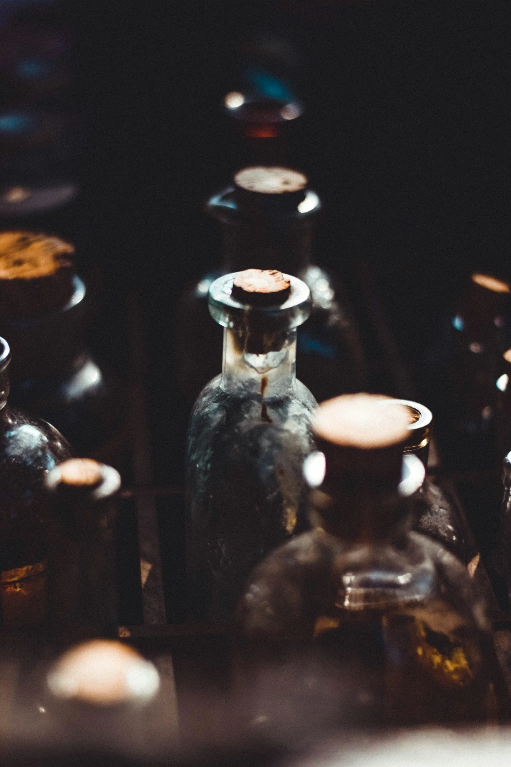 Une collection de fioles en verre anciennes et usées, avec des bouchons de liège, disposées dans un support noir dans un environnement sombre.