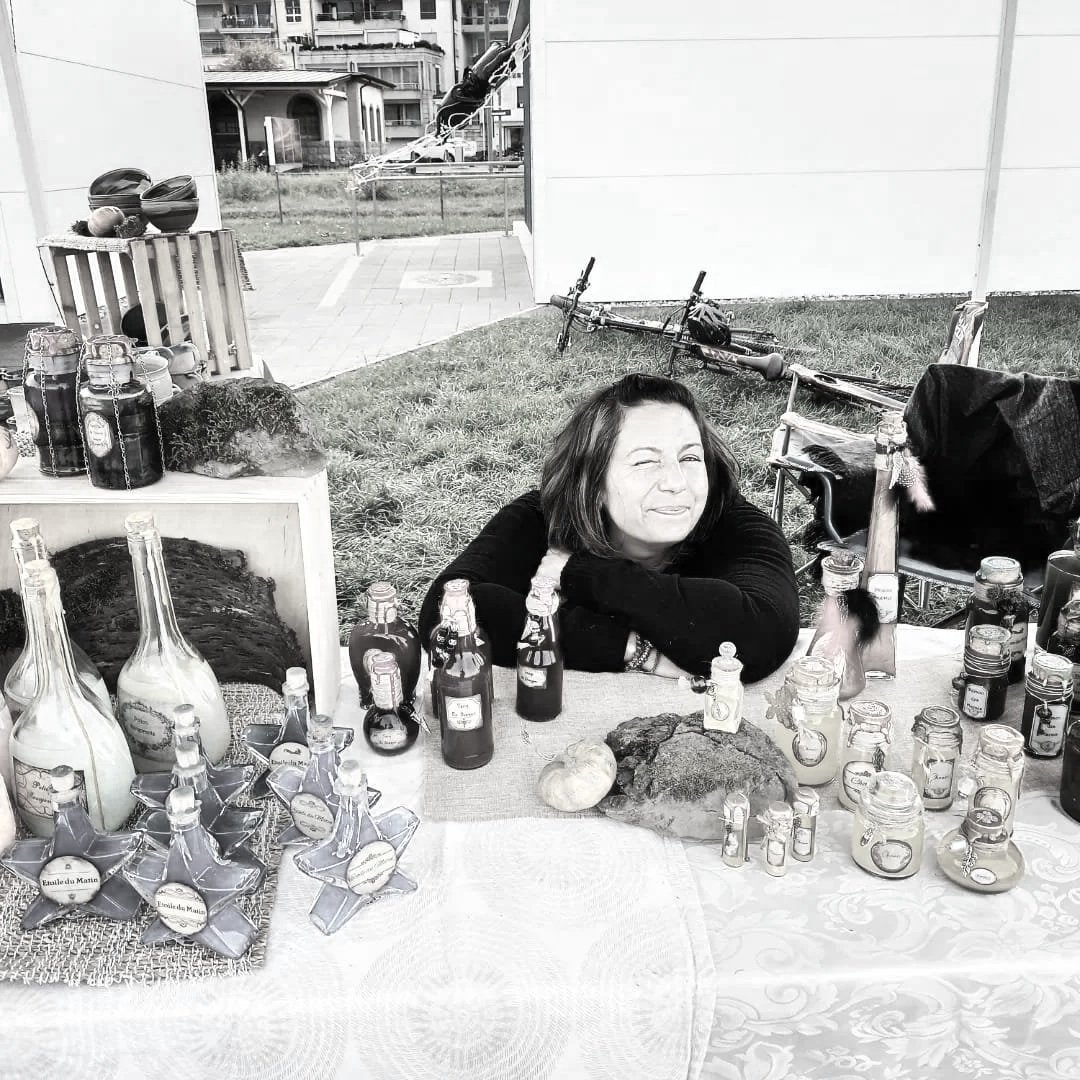 Une femme souriante  (Jessika Geoffroy) assise derrière un stand avec diverses bouteilles et petits bocaux, dans un espace extérieur. 