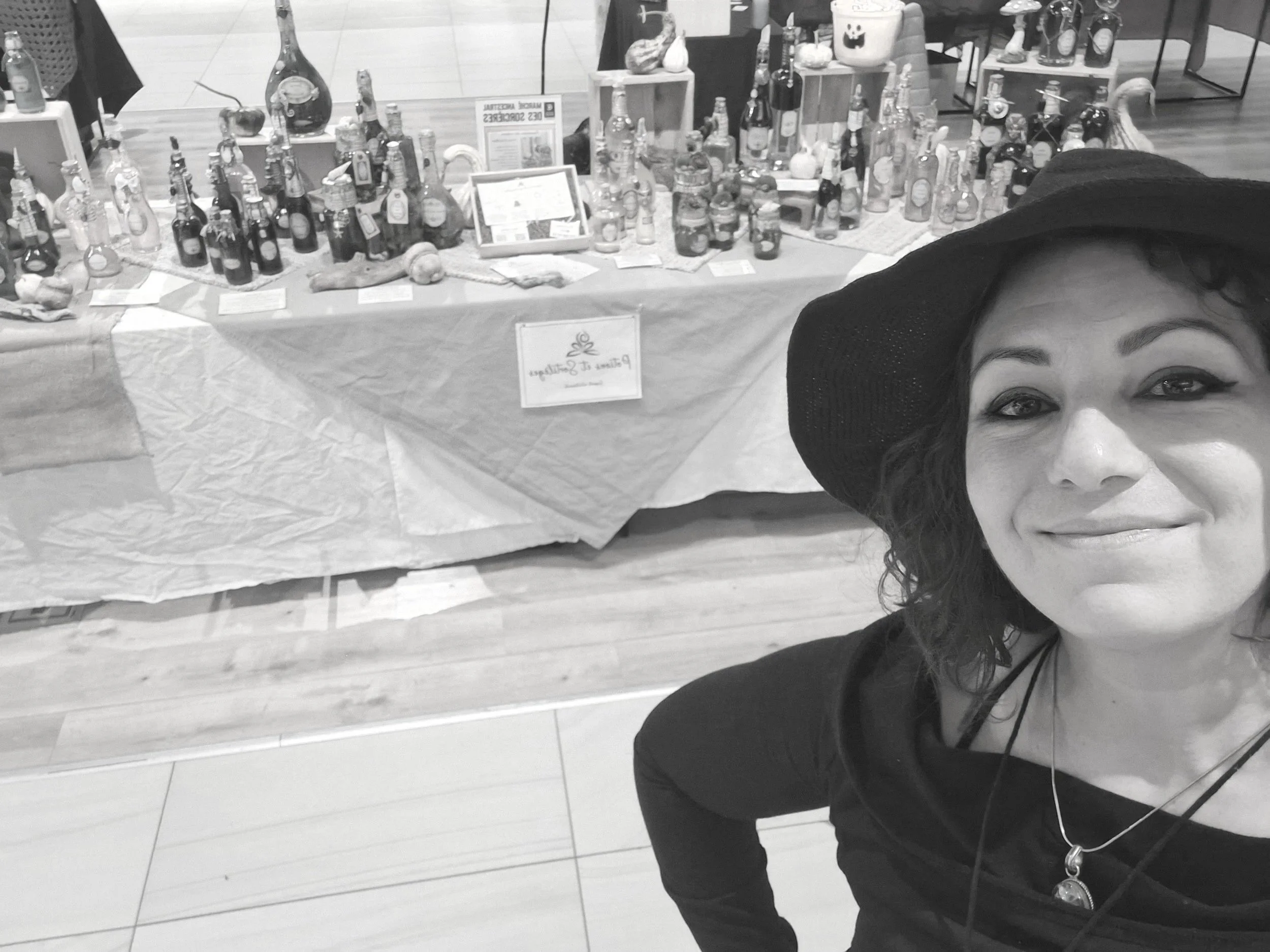 Une femme souriante  (Jessika Geoffroy)  portant un chapeau noir prend une selfie devant une table d'exposition remplie de bouteilles et d'objets divers.