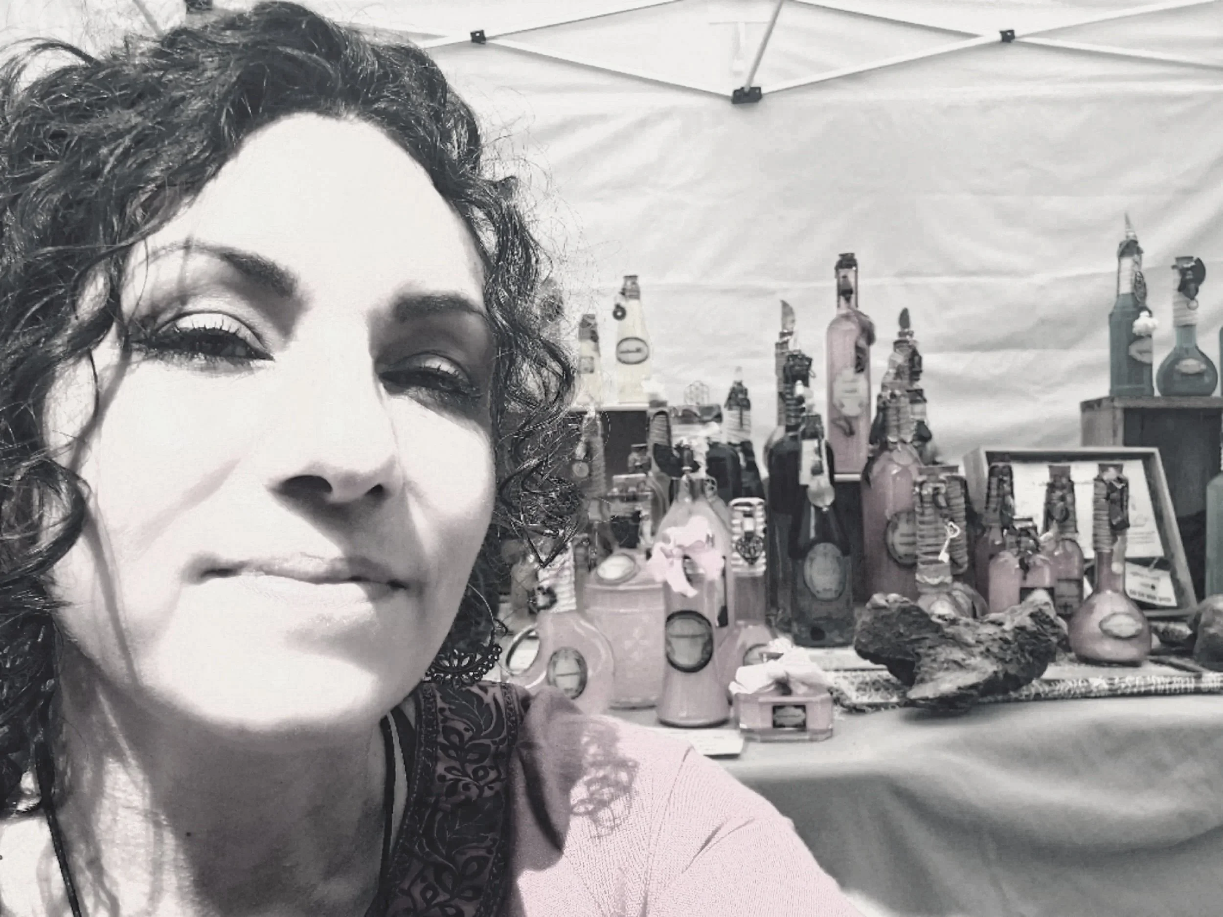 Une femme souriante avec des cheveux bouclés (Jessika Geoffroy) prend un selfie près d'une table avec des bouteilles en verre et des décorations artisanales, en plein air.