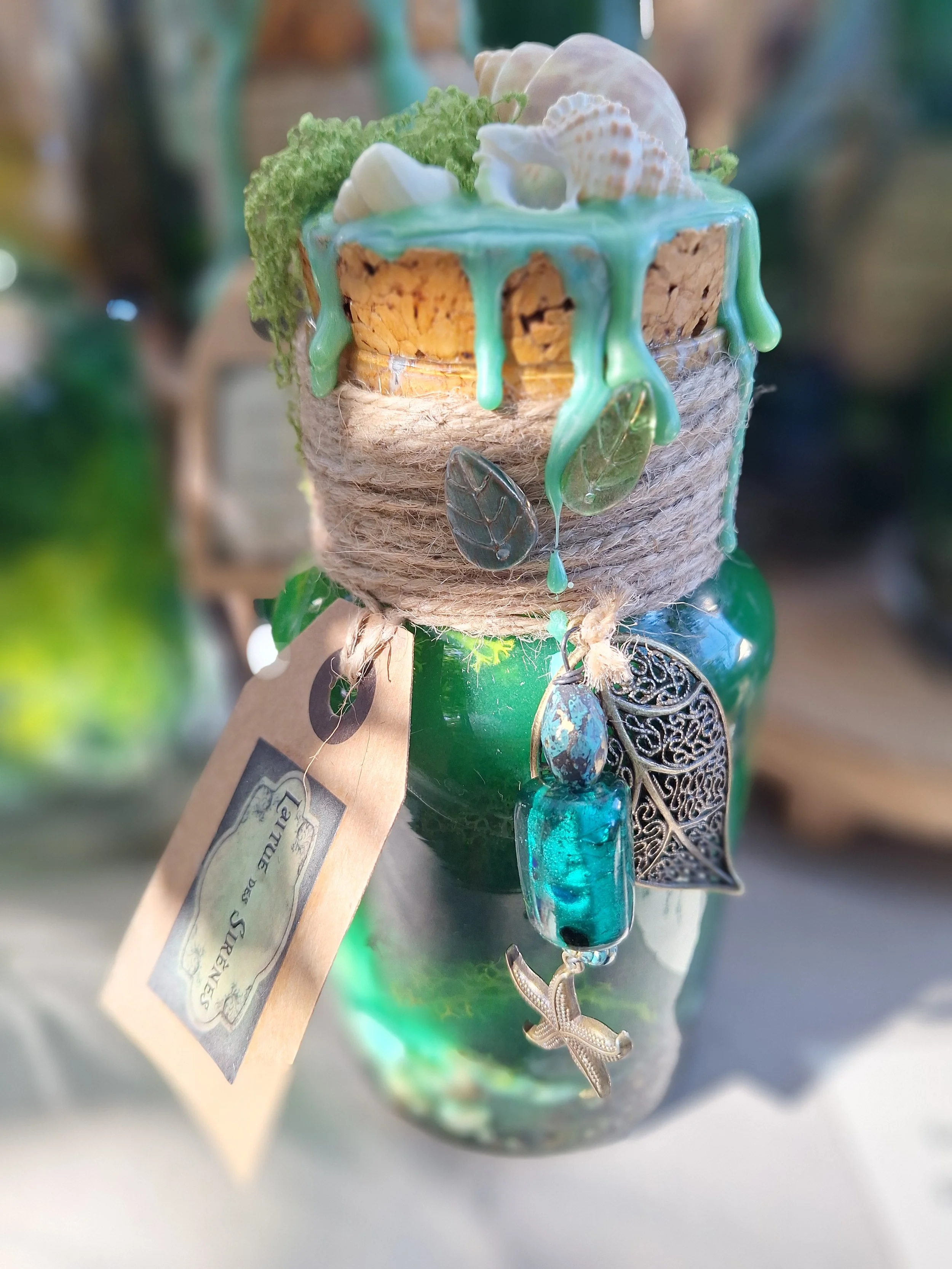 Une bouteille en verre verte décorée avec des pendentifs en forme de feuilles, une étoile de mer en métal, un clip de couleur turquoise et un petit oiseau en métal suspendus, avec un bouchon en liège recouvert de cire de couleur turquoise et ornée de