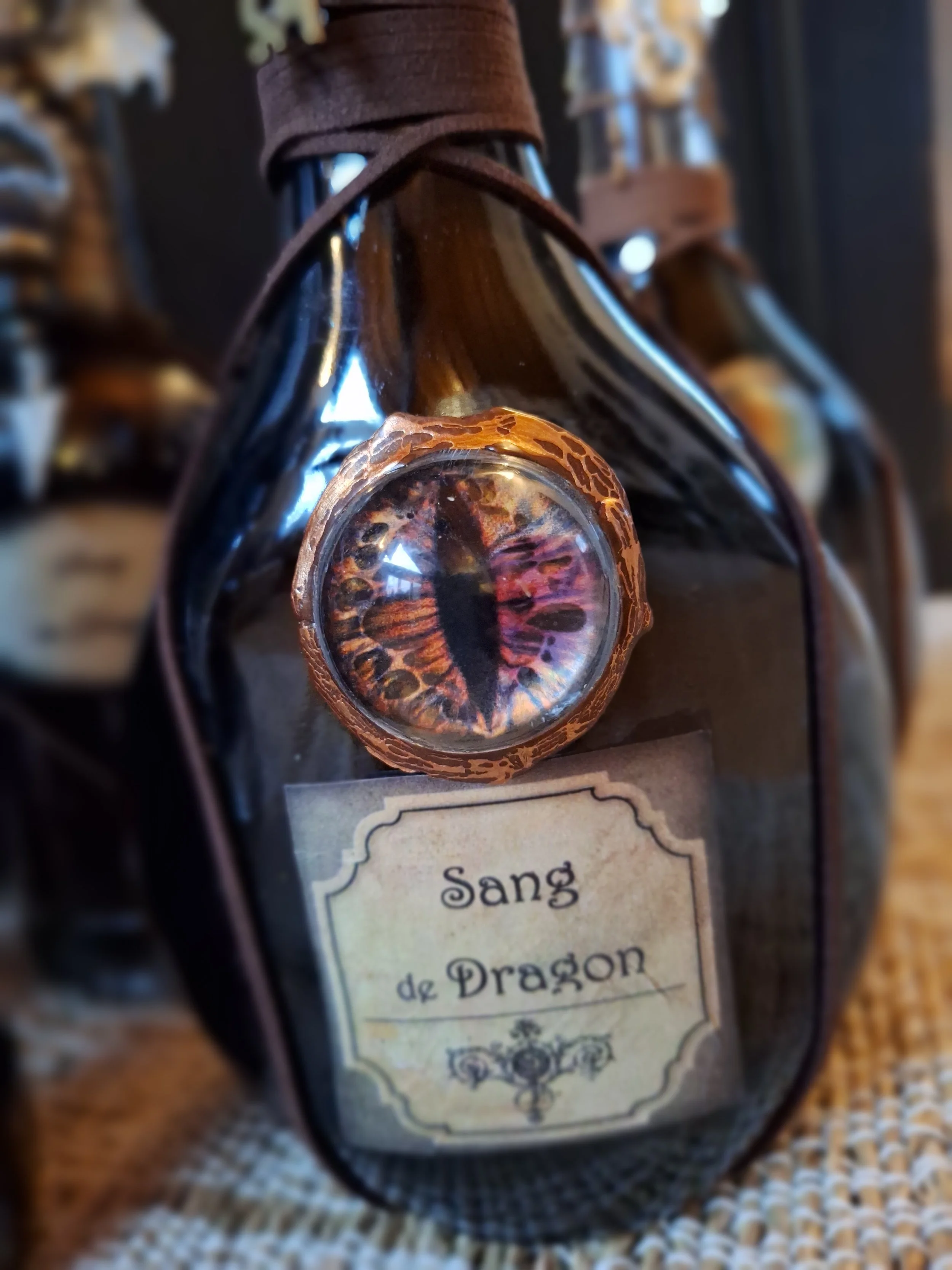 Une bouteille en verre sombre avec une étiquette portant l'inscription 'Sang de Dragon'. Sur la bouteille, il y a une capsule ou un élément décoratif représentant un œil de dragon avec une pupille verticale, évoquant un regard féroce et mystique.