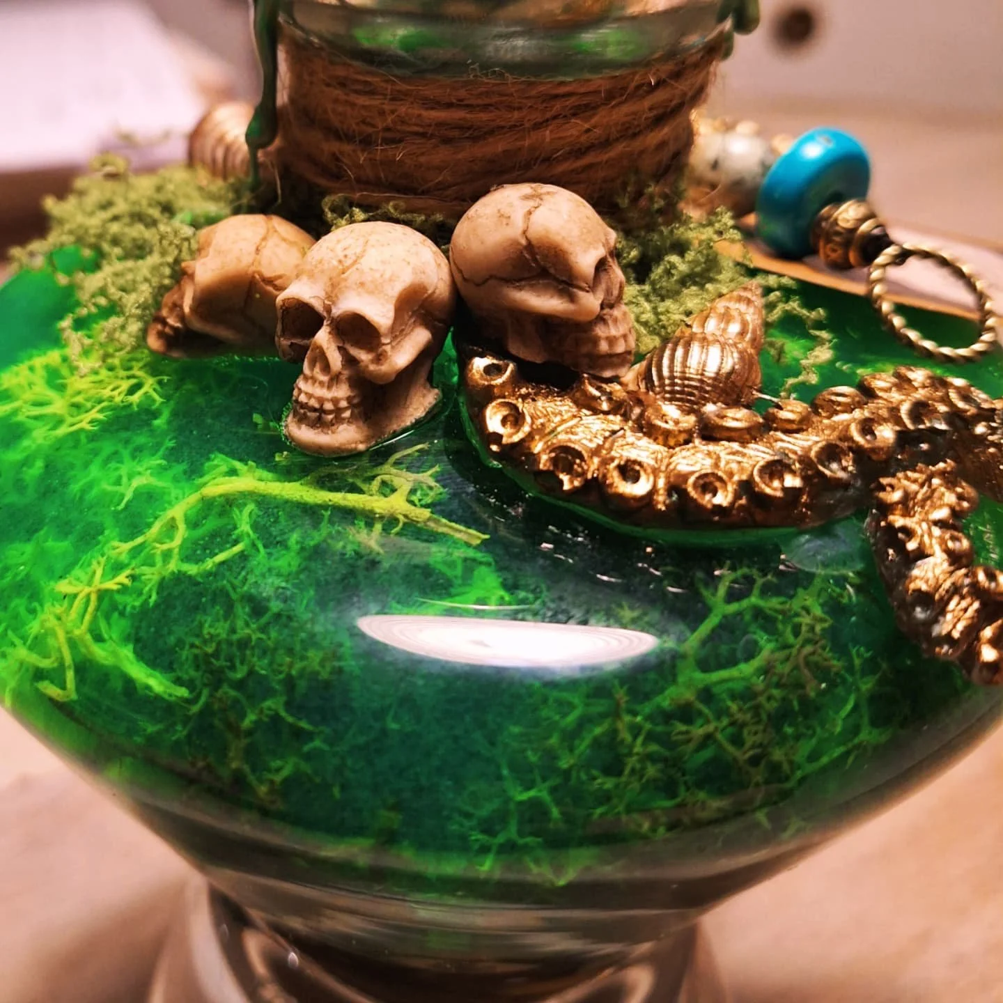 Carafe en verre décorée avec des petits crânes, un poulpe en métal doré, des bijoux et des éléments verts sous une surface en résine transparente