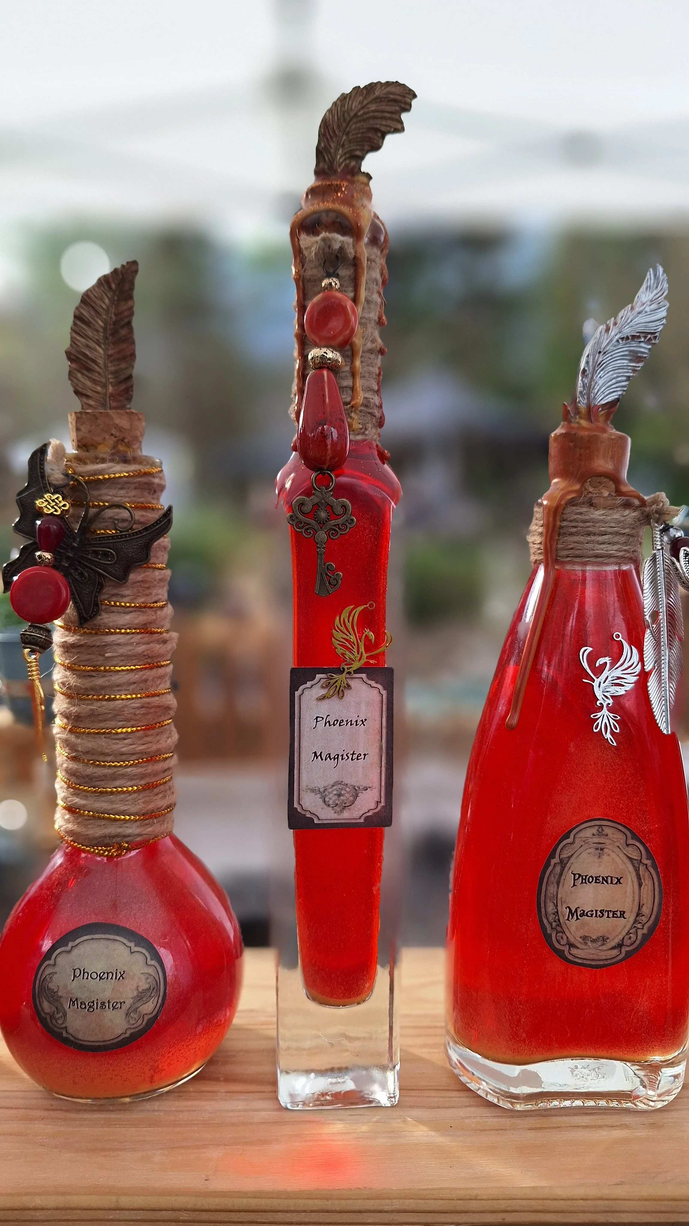 Trois fioles rouges avec des plumes, des perles et des étiquettes portant la mention 'Phoenix Magister', posés sur une surface en bois.