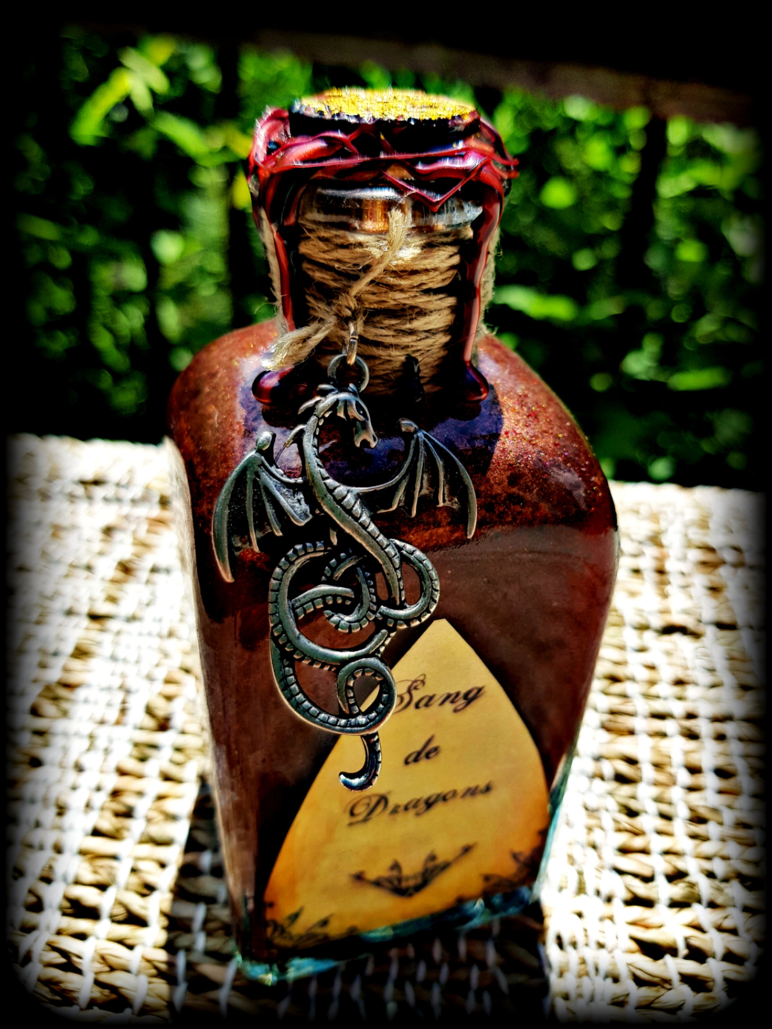 Bouteille en verre décorée avec un pendentif dragon, étiquette 'Sang de Dragon', bouchon en lit de corde, en extérieur sur une surface en osier.