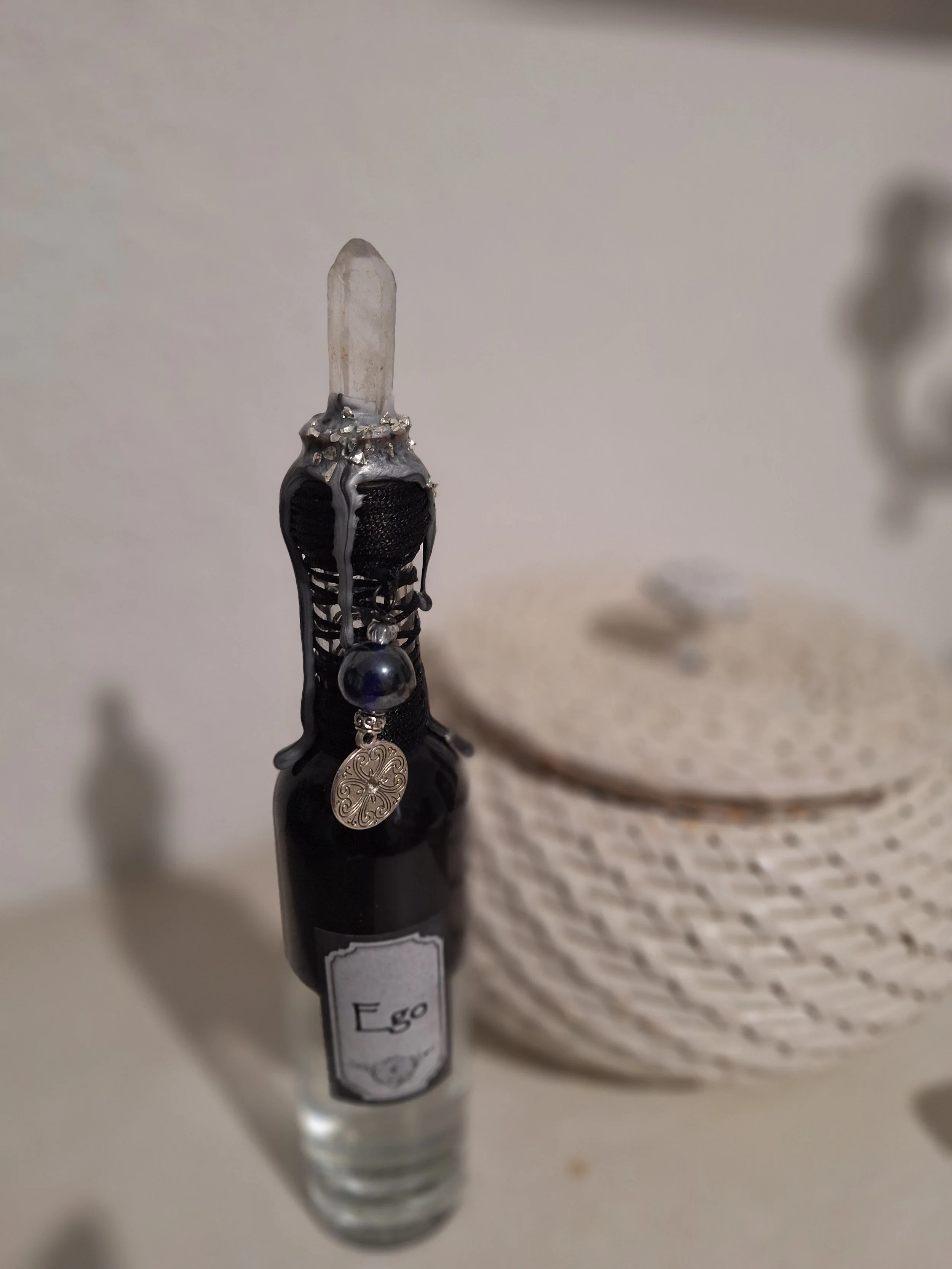 Une bouteille allongée avec un bouchon décoré de bijoux, un cristal en haut, et une étiquette portant le mot 'Ego'.