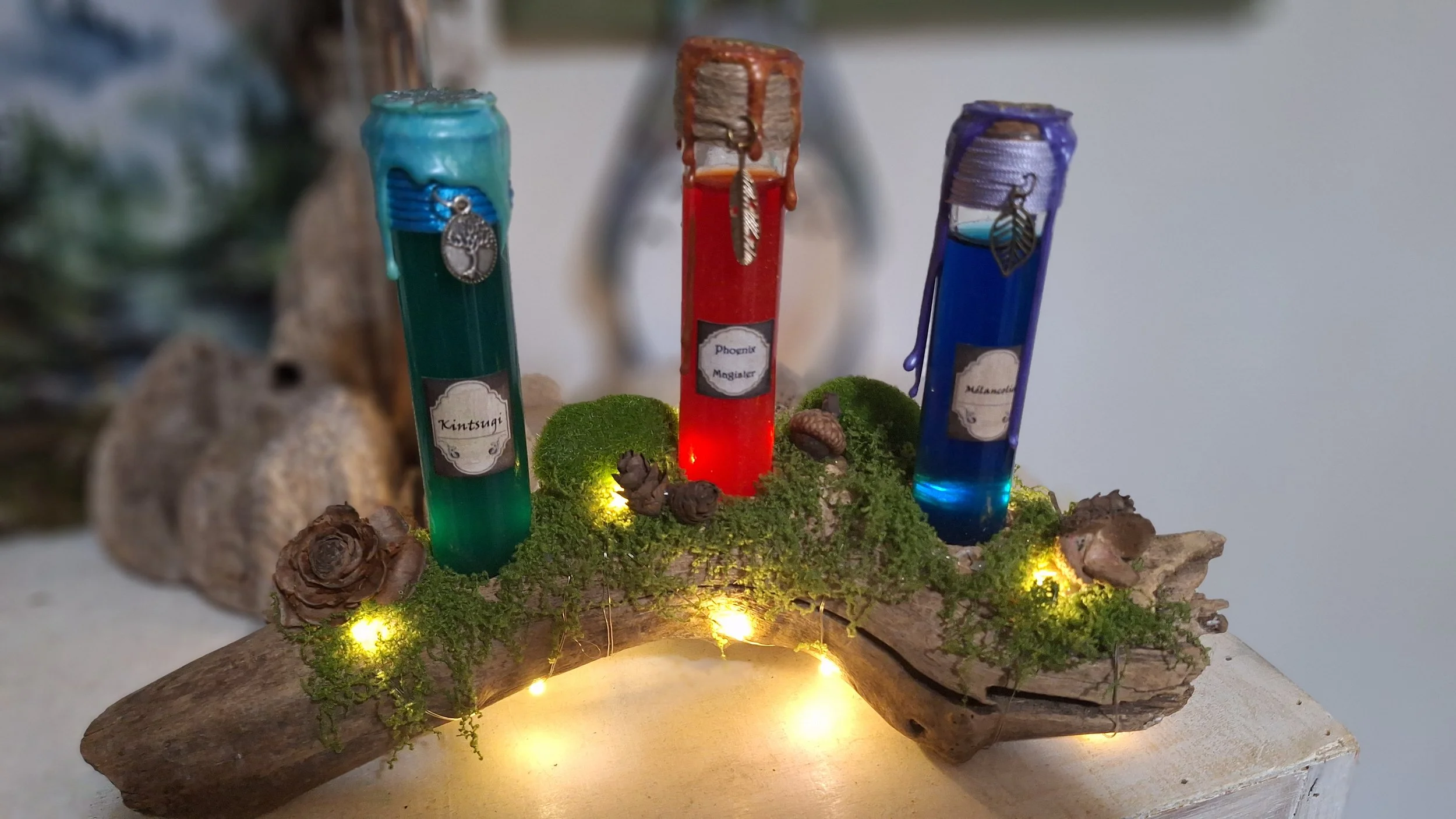 Trois fioles colorées (verte, rouge et bleue) avec des étiquettes portant les noms 'Kintsugi', 'Phoenix Mangister' et 'Mélanclia', posées sur un morceau de bois recouvert de mousse, avec de petites lumières et des éléments naturels décoratifs autour.