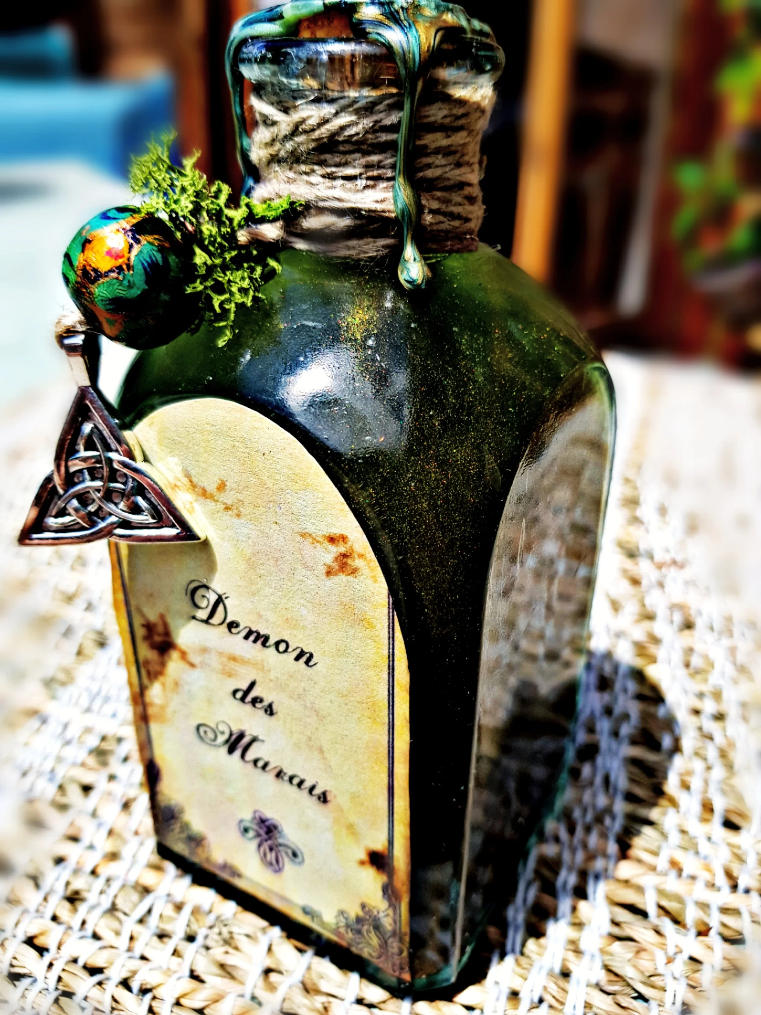 Une bouteille en verre avec une étiquette portant l'inscription « Démon des Marais ». La bouteille est décorée avec une boule colorée et des éléments en métal en forme de triquetra, posée sur un tapis en osier.