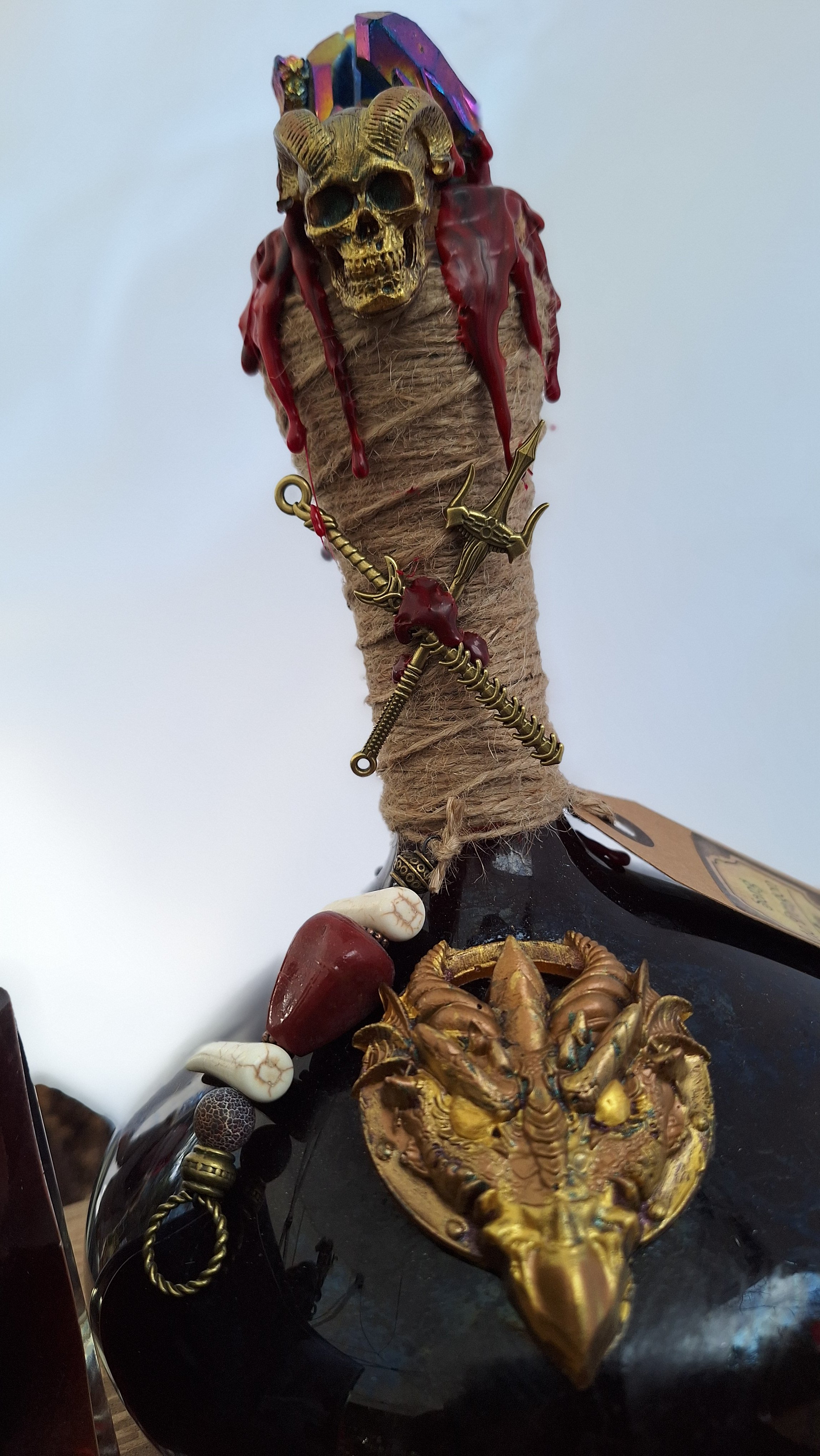 Une carafe en verre décorée d'accessoires sombres et mystérieux, notamment un crâne doré, des pendentifs en forme de dents, et des éléments en métal doré, créant une apparence gothique.