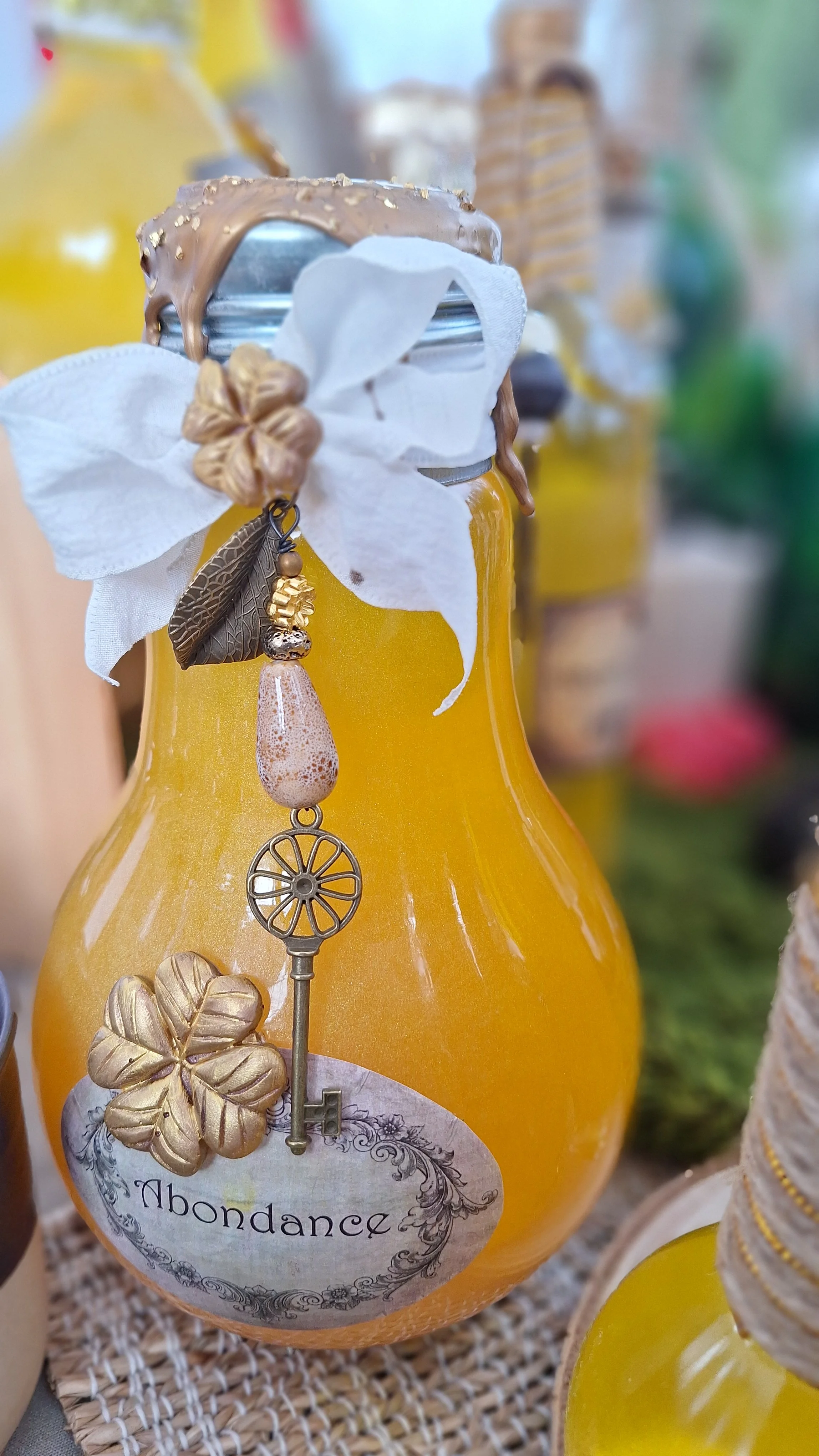 Bouteille en verre avec étiquette 'Abondance', décorée d'une fausse fleur blanche, d'une feuille en métal, et d'un pendentif avec une clé. La bouteille est jaune/orangée avec une étiquette vintage.