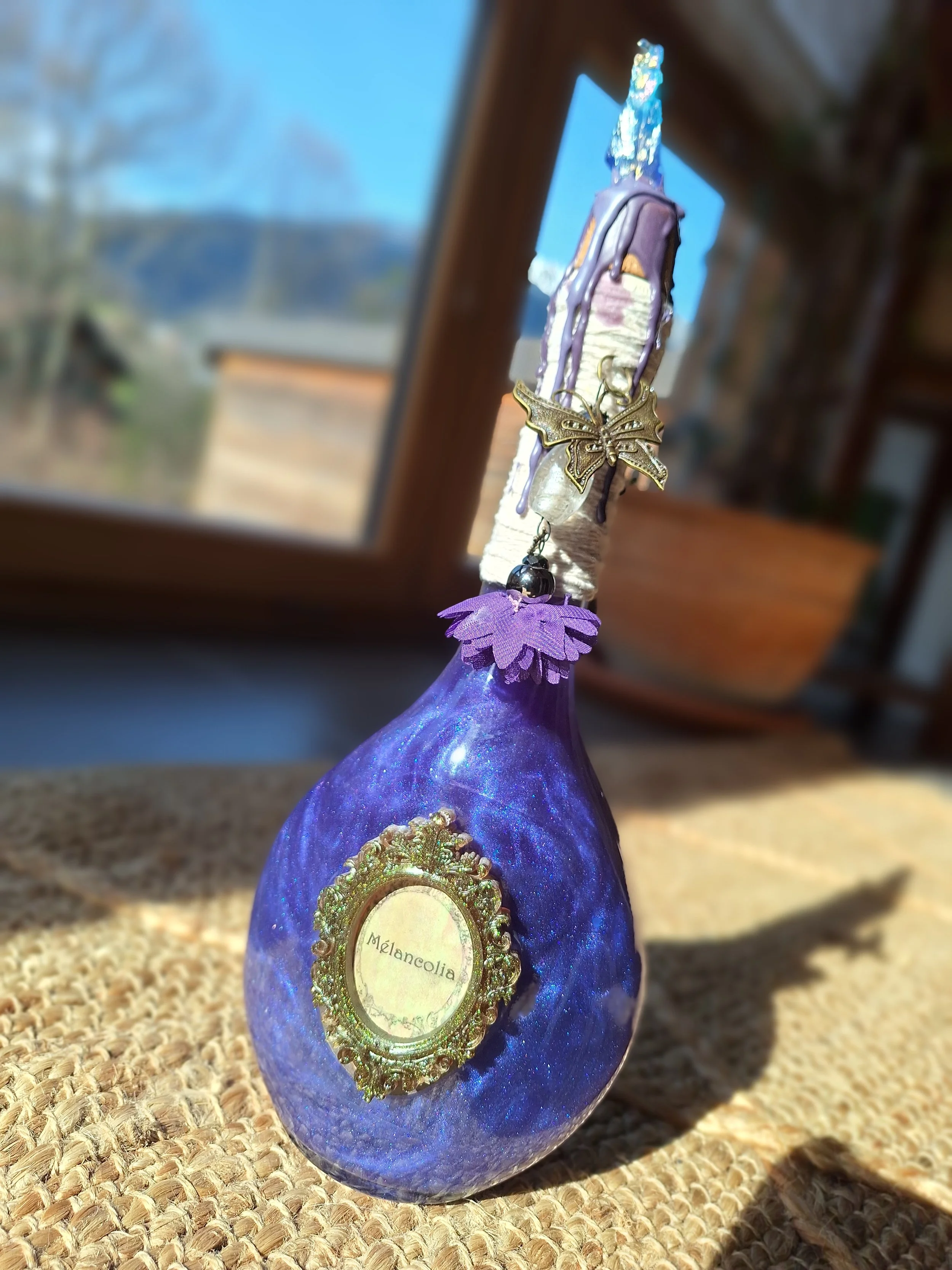 Une bouteille décorative violette avec une étiquette 'Mélancolia', ornée d'une breloque papillon et d'une décoration en tissu violet, posée sur une surface en osier près d'une fenêtre.