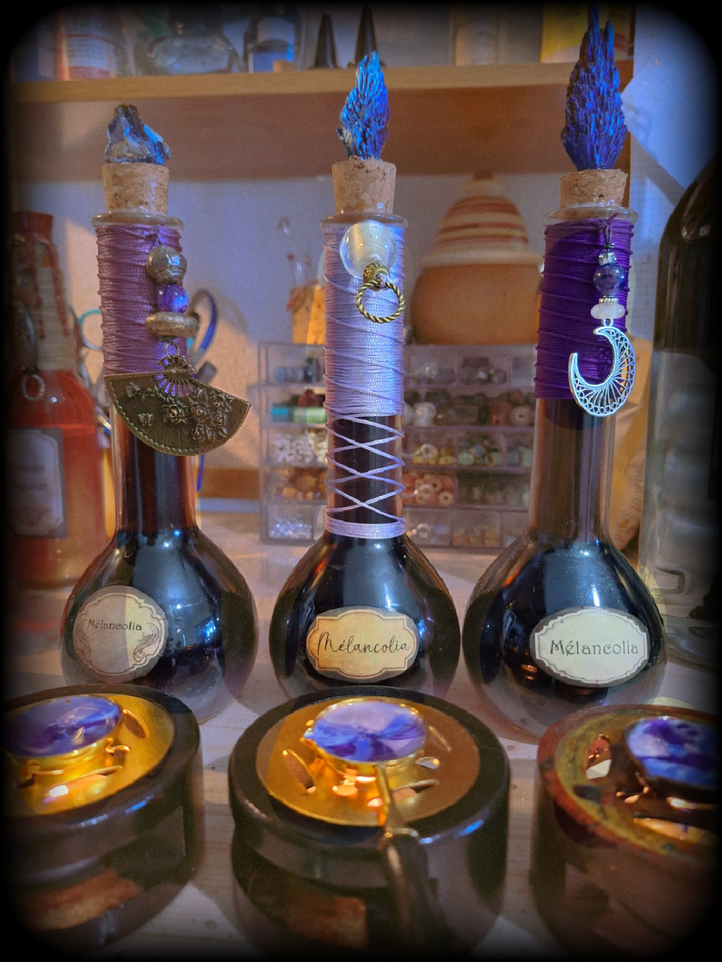 Trois fioles décoratives contenant un liquide violet, avec des bouchons en liège, décorés avec des rubans violets et des pendentifs, et des bougies allumées en dessous.