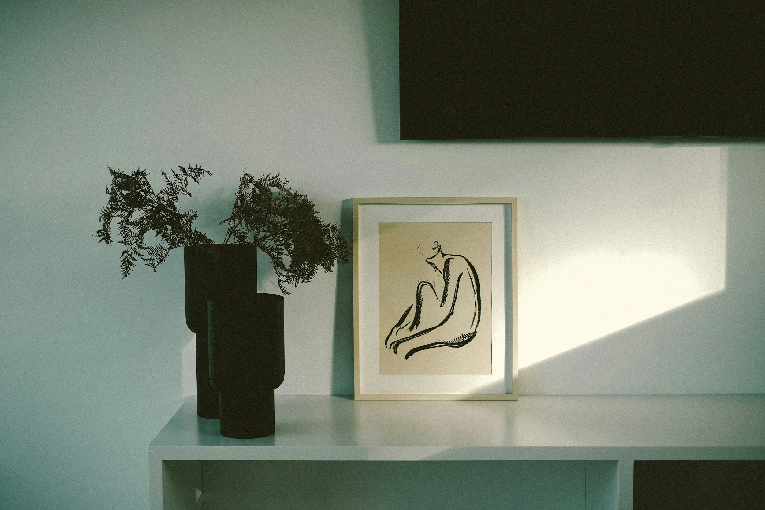A framed minimalist line drawing of a seated person with legs crossed, placed on a white surface next to a black vase with fern-like plant, against a light wall with sunlight creating a shadow.