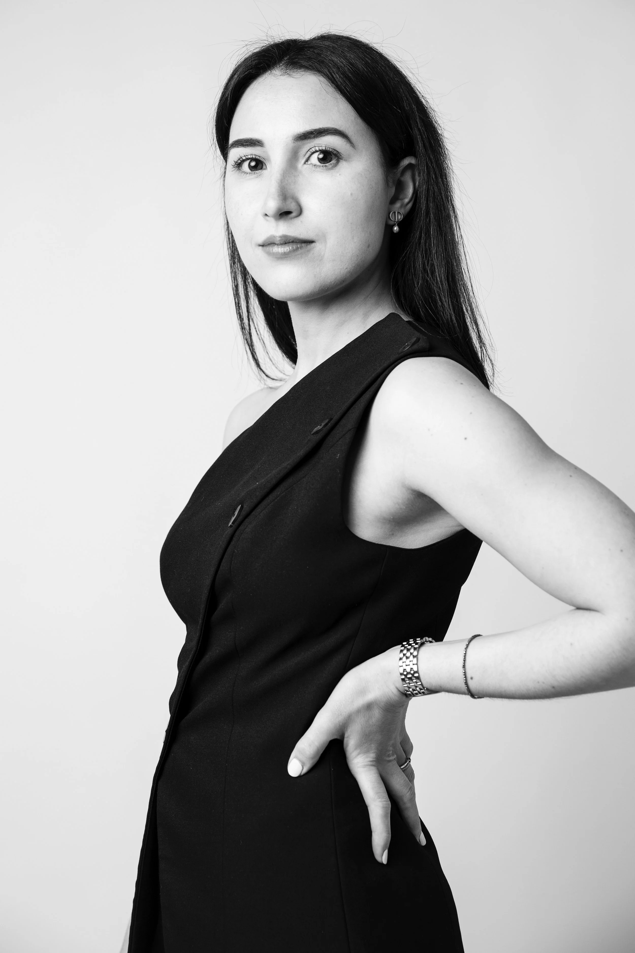 Une femme en noir et blanc, portant une robe noire, avec une montre et des bracelets, se tenant de profil avec une main sur la hanche, regardant vers l'objectif.