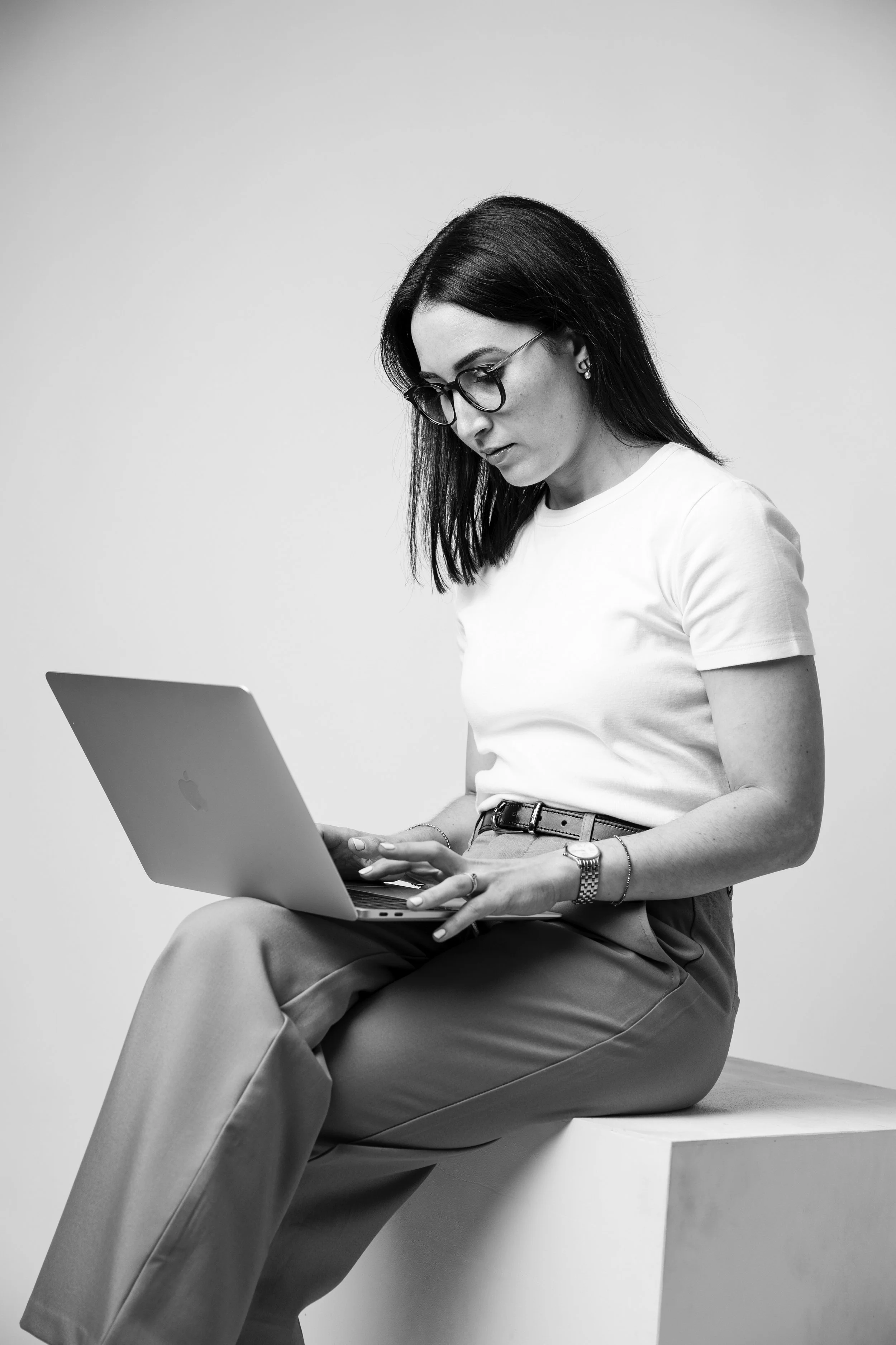 Une femme élégante portant des lunettes, un t-shirt blanc et un pantalon en tissu clair, assise sur un siège en bois et utilisant un ordinateur portable.