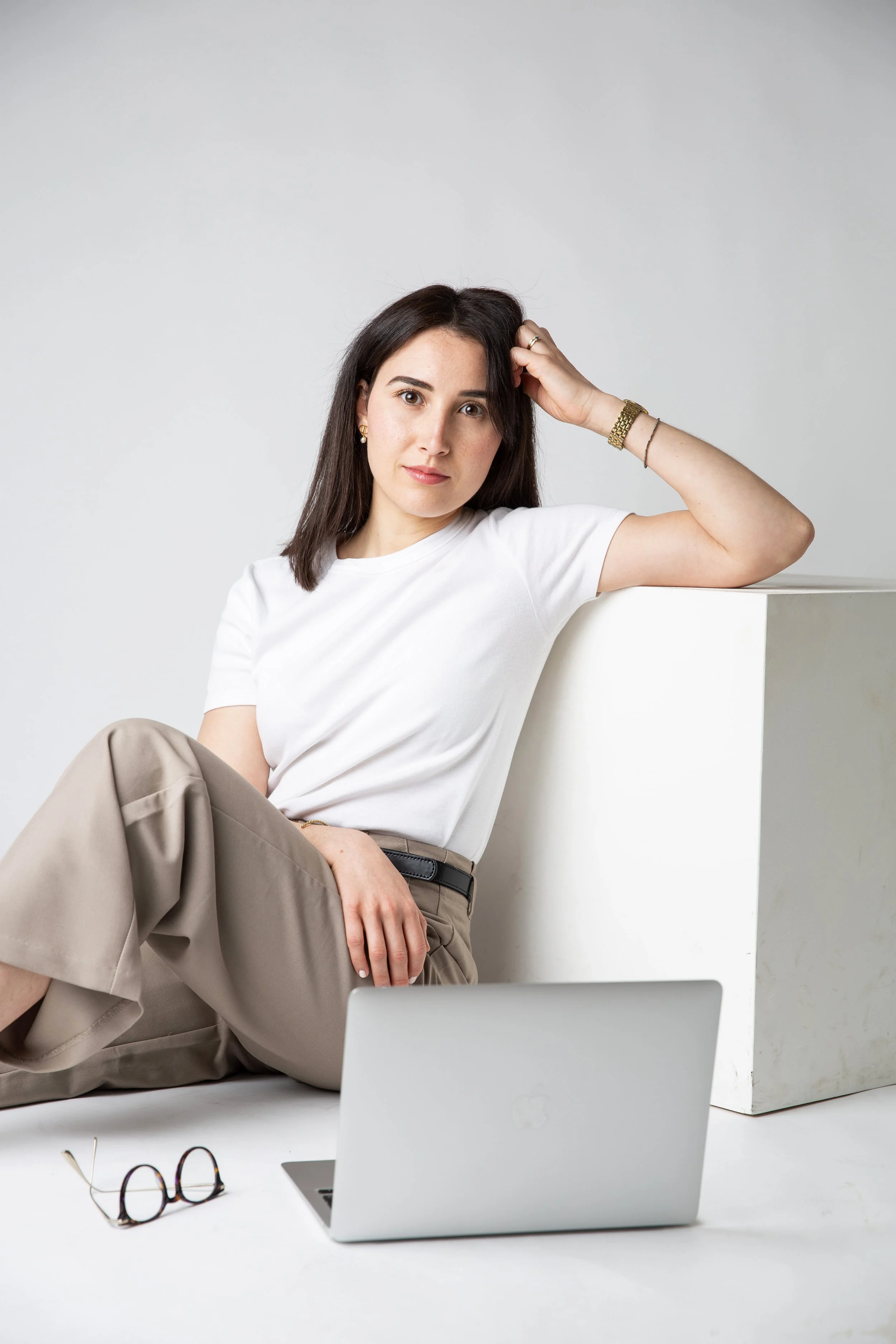 Une jeune femme assise devant un ordinateur portable blanc, portant un t-shirt blanc et un pantalon beige ample, avec des lunettes à côté d'elle, dans un environnement minimaliste moderne.