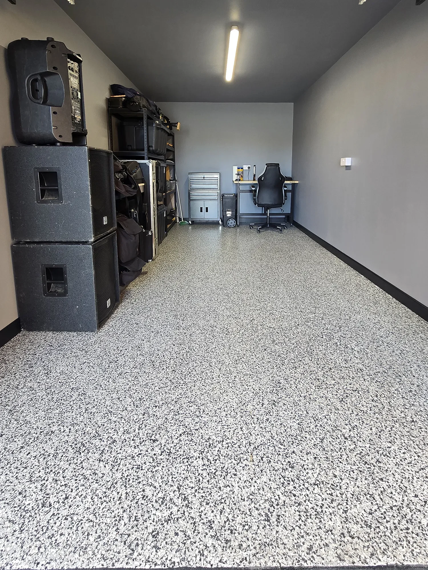 Garage with a speckled resin flooring, and minimal furniture including a desk with office supplies, a black office chair, a metal storage cabinet, and black shelving with equipment and bags, illuminated by a ceiling light.