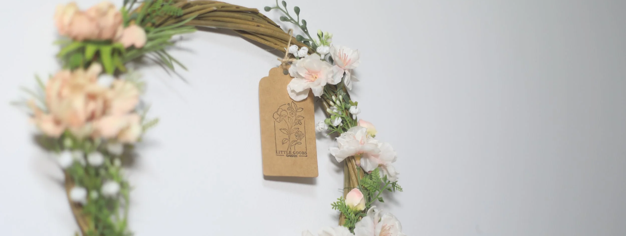 A floral wreath with pink and white flowers and green foliage hanging on a white wall. A brown tag with a floral illustration and the text 'Little Goobs Garden' is attached to the wreath.