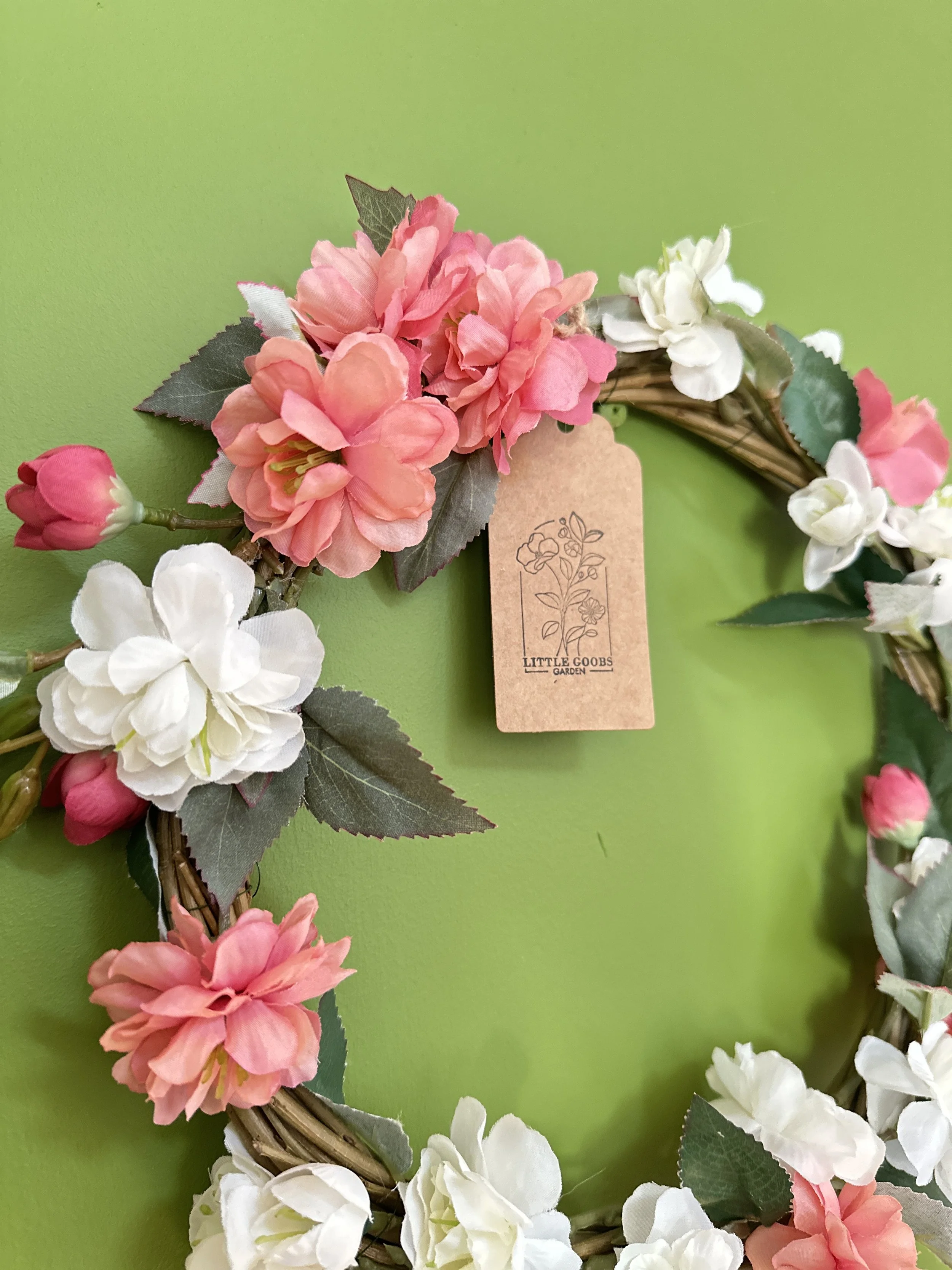A floral wreath with pink, white, and peach fabric flowers, including green leaves, hanging against a bright green background, with a brown tag reading 'Little Goobs Garden' attached to it.