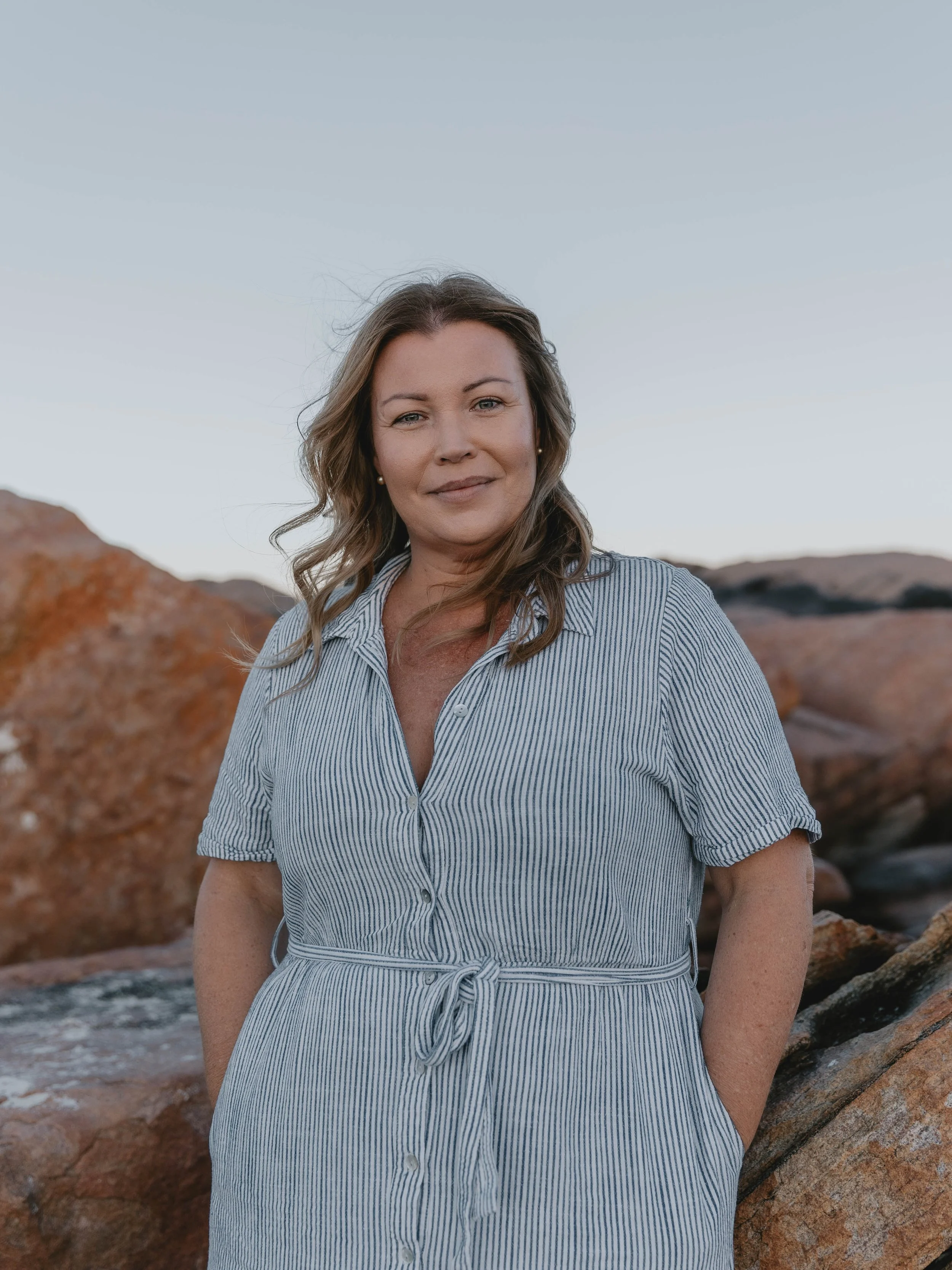 A woman with shoulder-length wavy hair, wearing a blue and white striped dress, standing outdoors near rocks with a clear sky in the background.