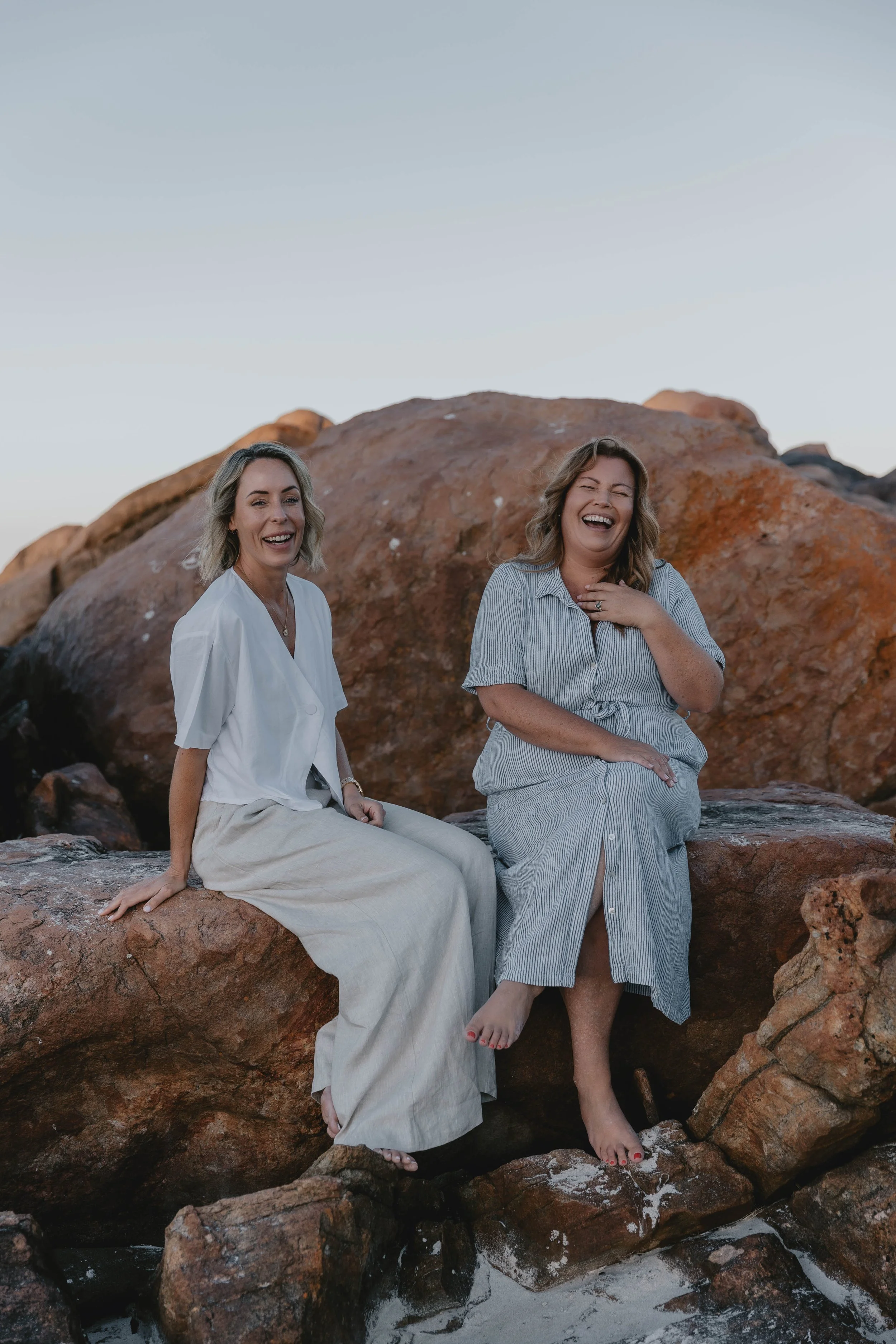 Two women sitting on rocks at the beach, smiling and laughing, with the sky in the background.