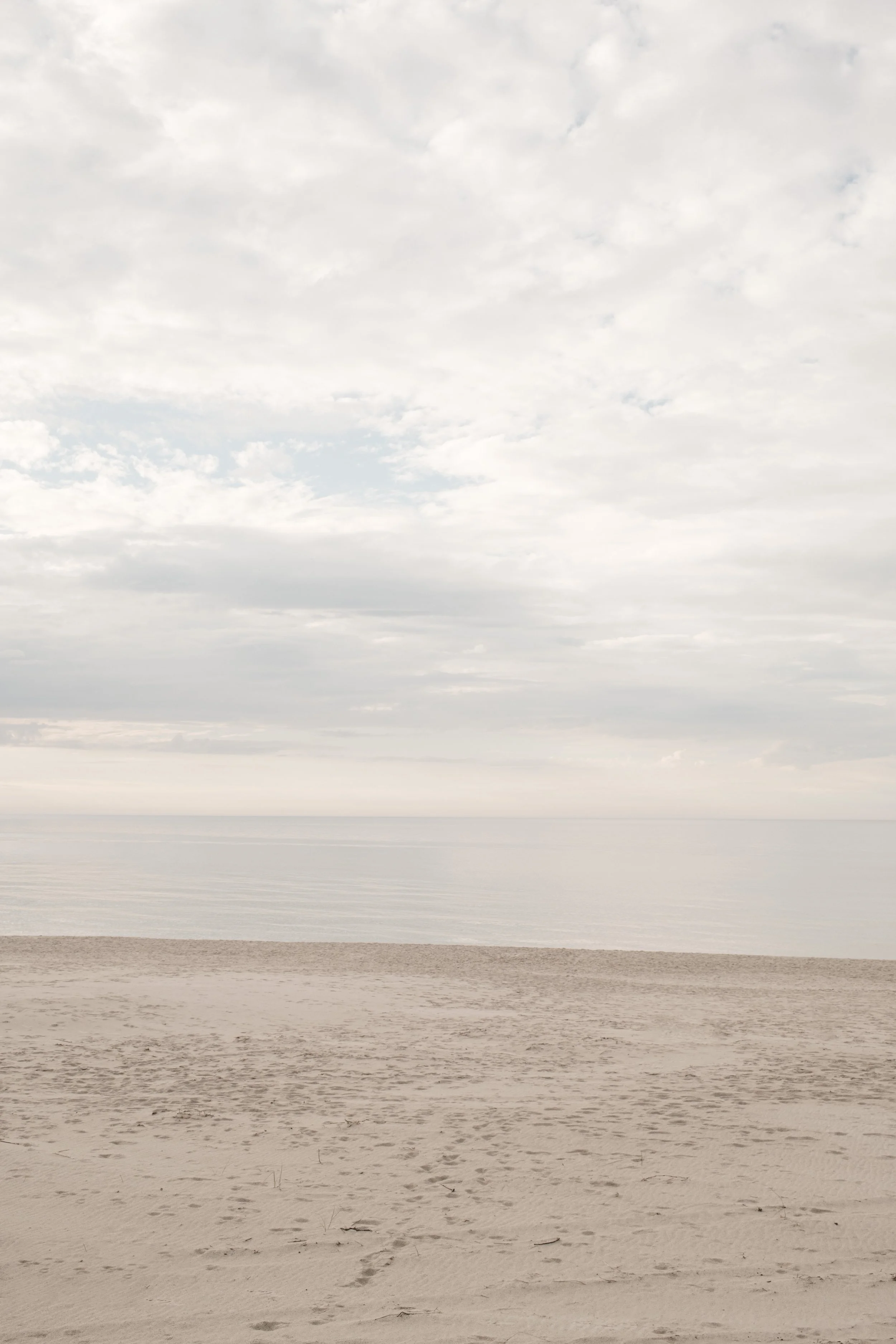 Beach scene with sand in the foreground, calm ocean water in the middle, and a cloudy sky above.