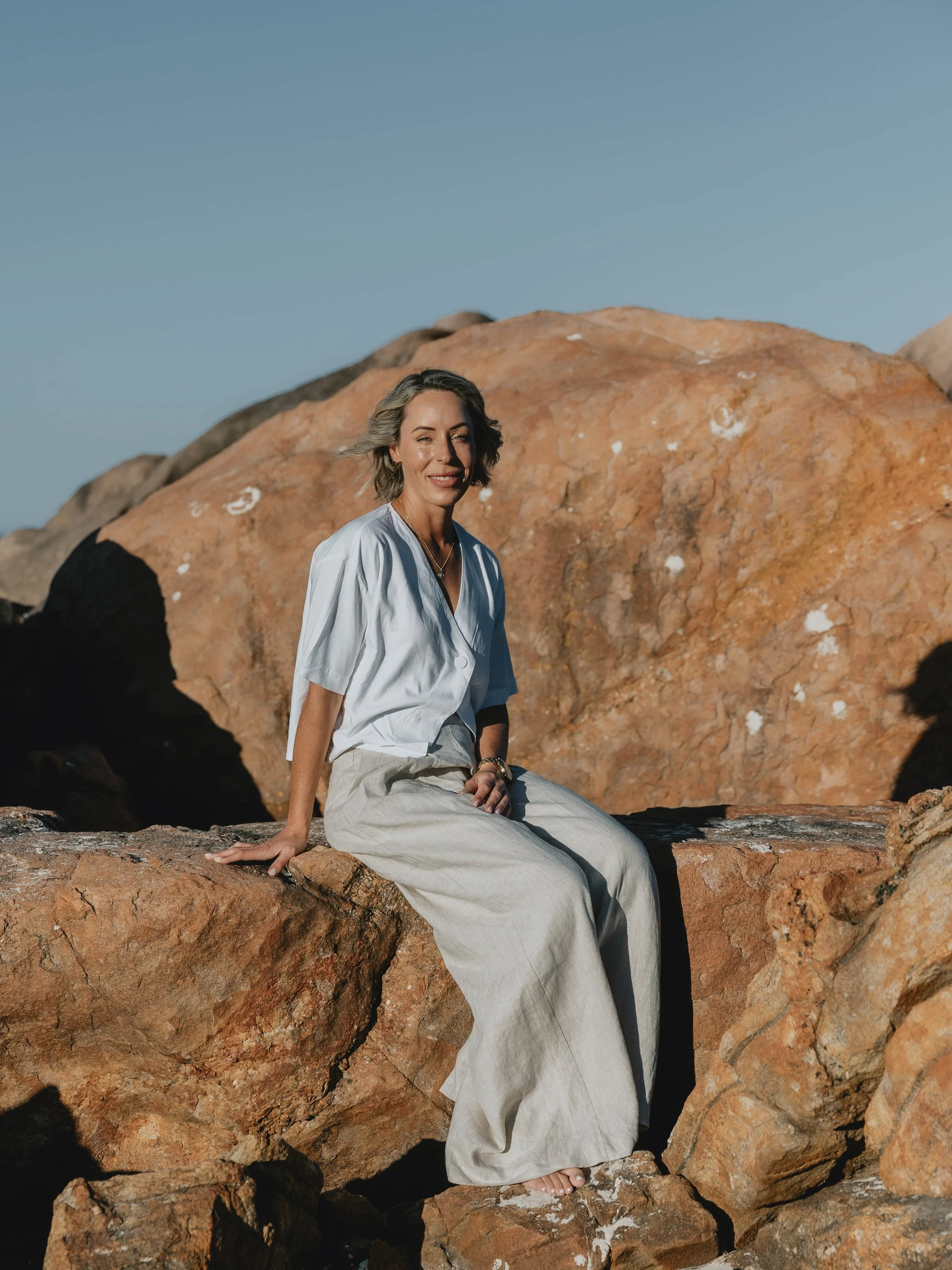 A woman with short blonde hair sitting on large rocks, wearing a white blouse and beige pants, with a clear blue sky in the background.
