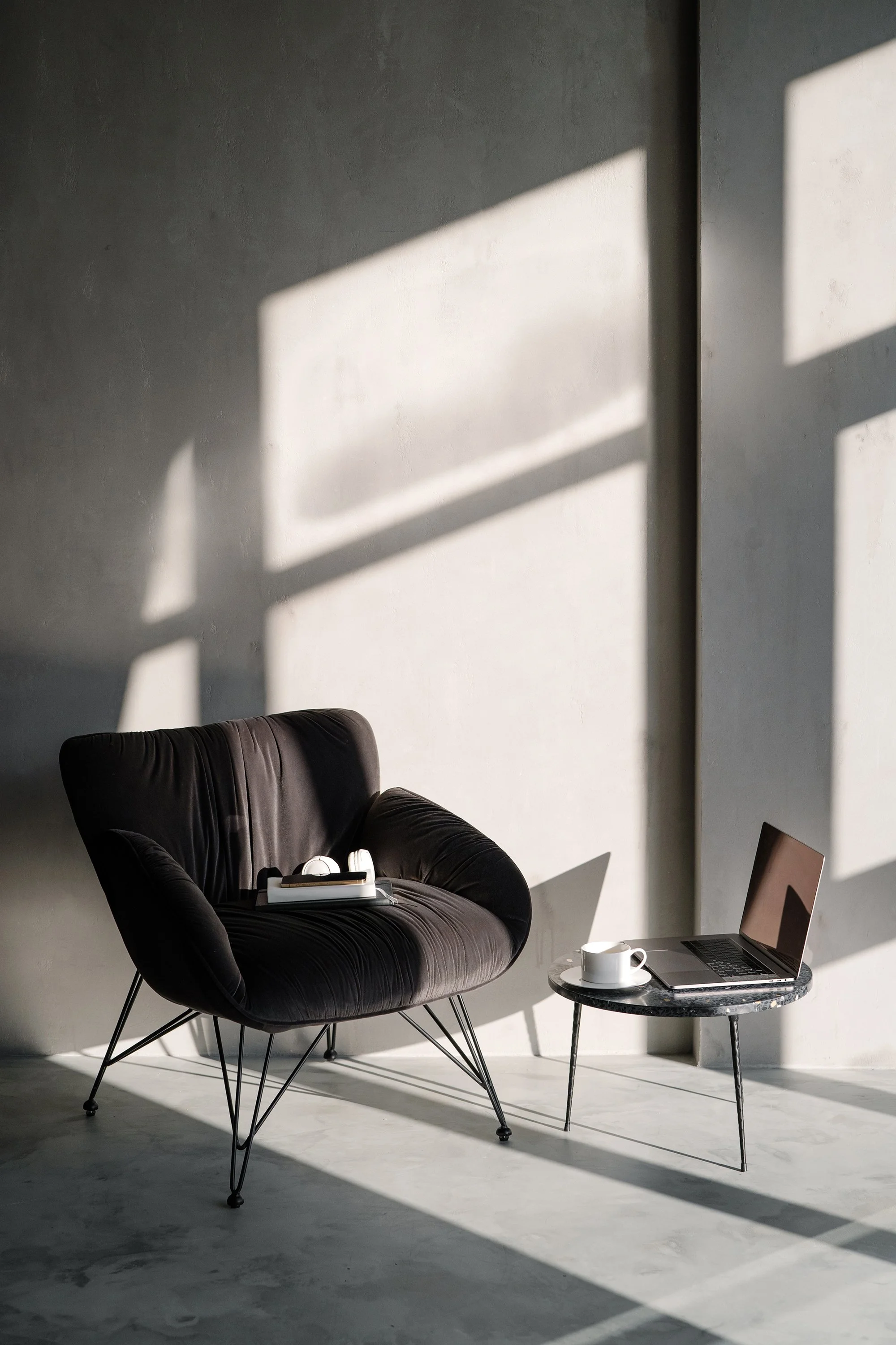 A black armchair with metal legs positioned next to a small round marble table. On the table, there is a white cup and a laptop. Shadows of window frames cast geometrical patterns on a plain, light-colored wall, indicating sunlight coming through a window.