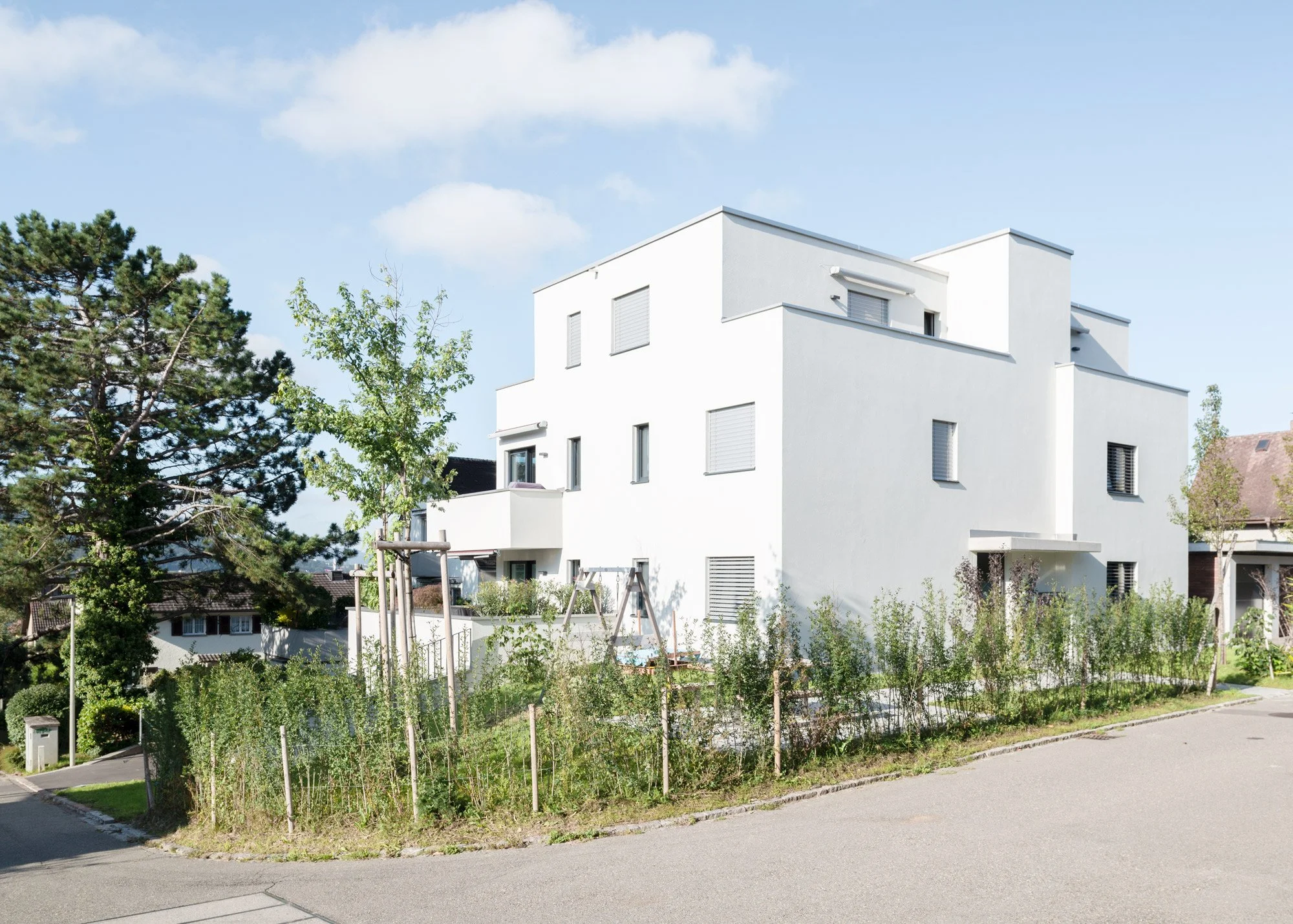 Modern weisses Mehrfamilienhaus in Wallisellen ZH mit mehreren Etagen und kleinen Balkonen, umgeben von einem Garten mit Blumen und Bäumen, bei schönem Himmel.