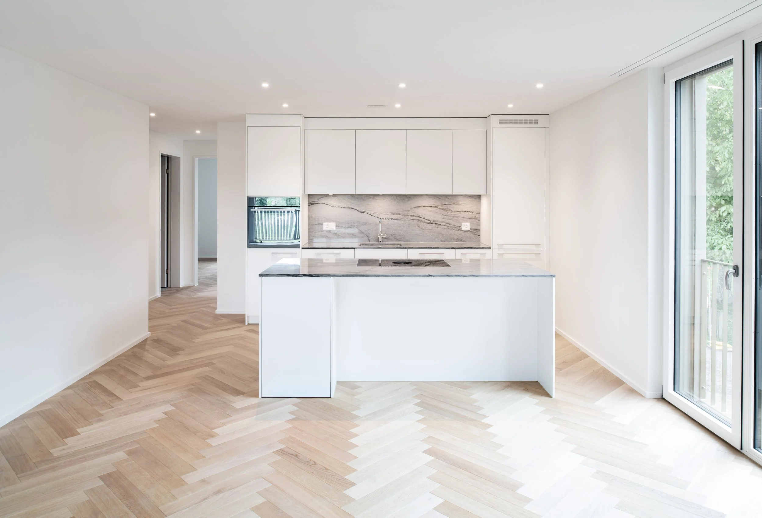 Moderne leere Wohnungsküche mit weissen Schränken, Markenschleppplatte, Kochstudio, Holzboden und grossen Fenstern, die nach draussen führen.