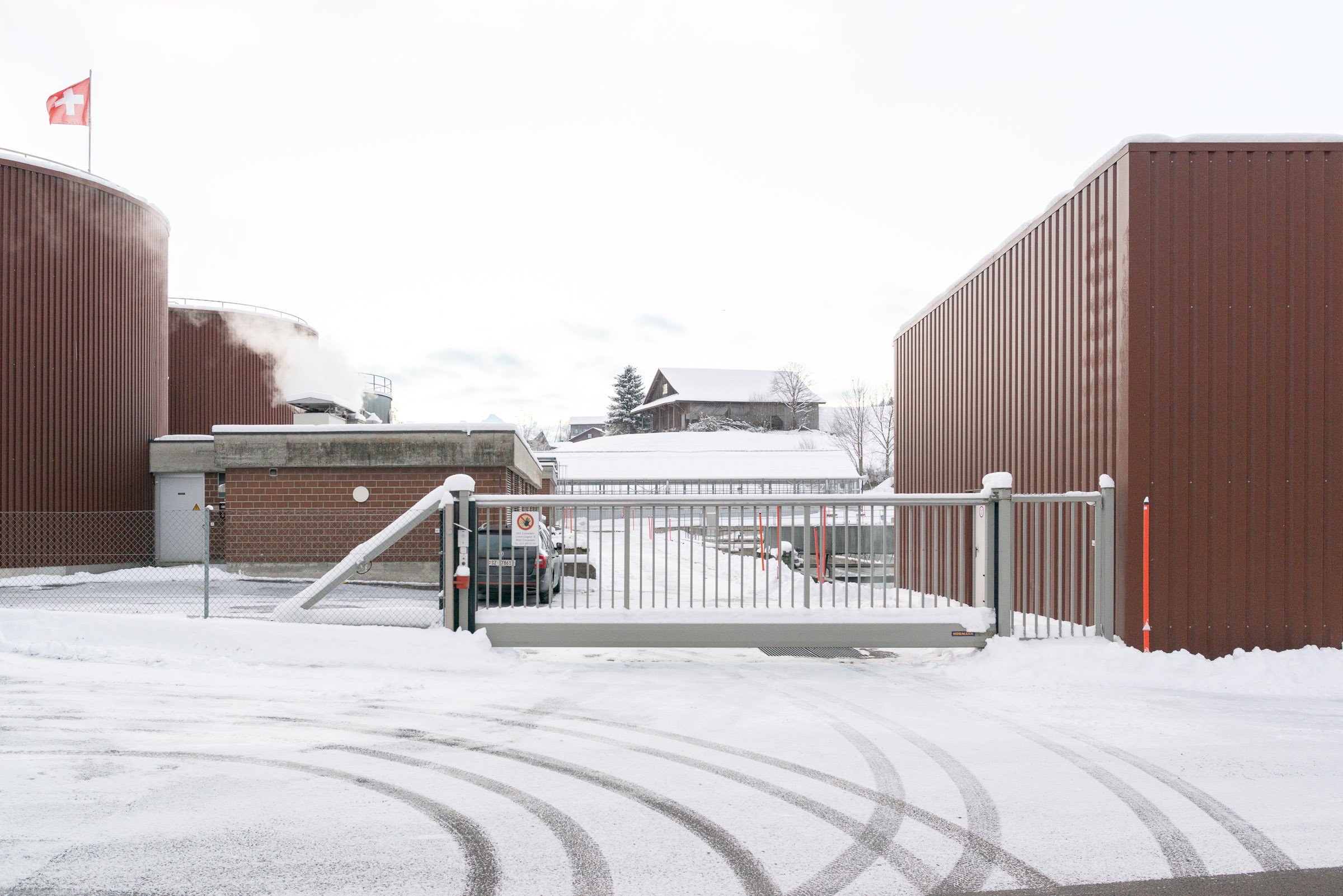 Schneebedeckte Strasse in einem industriellen Bereich mit hohen rotbraunen Gebäuden und Schweizer Flagge im Hintergrund.