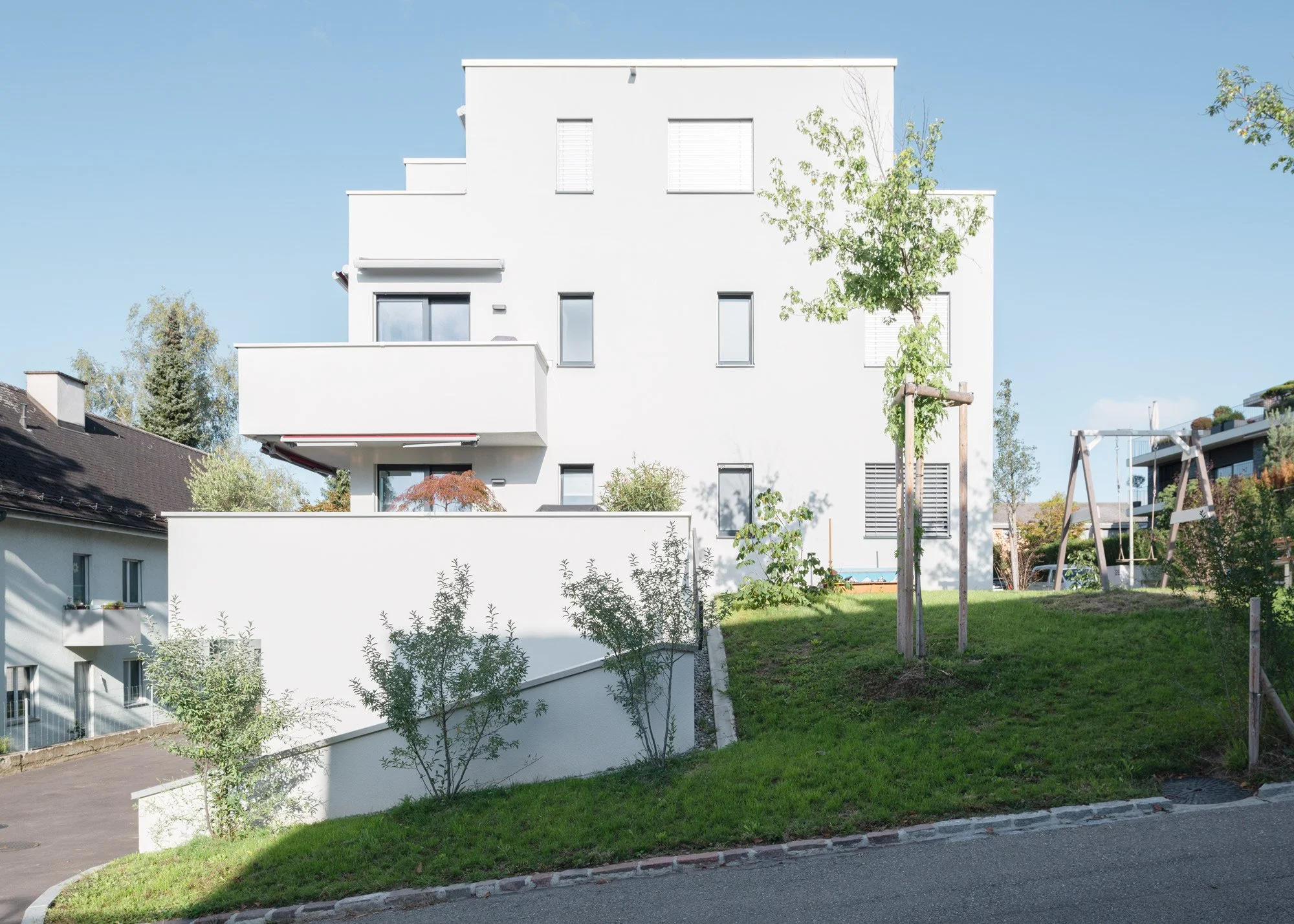 Modern weisses Mehrfamilienhaus mit mehreren Etagen, Balkonen und kleinen Gärten im Vordergrund. Es gibt Bäume und einen Spielplatz im Gartenbereich, ein Himmel ist sichtbar.