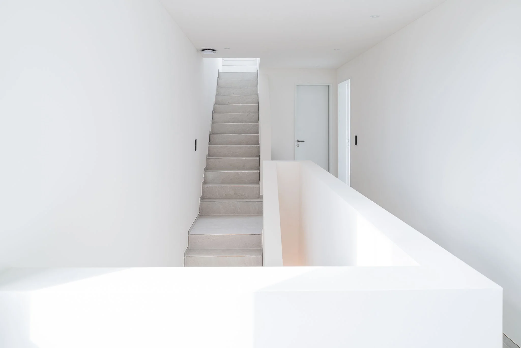 Innenaufnahme eines minimalistisch gestalteten Flurs mit weissen Wänden, einer Treppe auf der linken Seite und einer weissen Tür auf der rechten Seite.