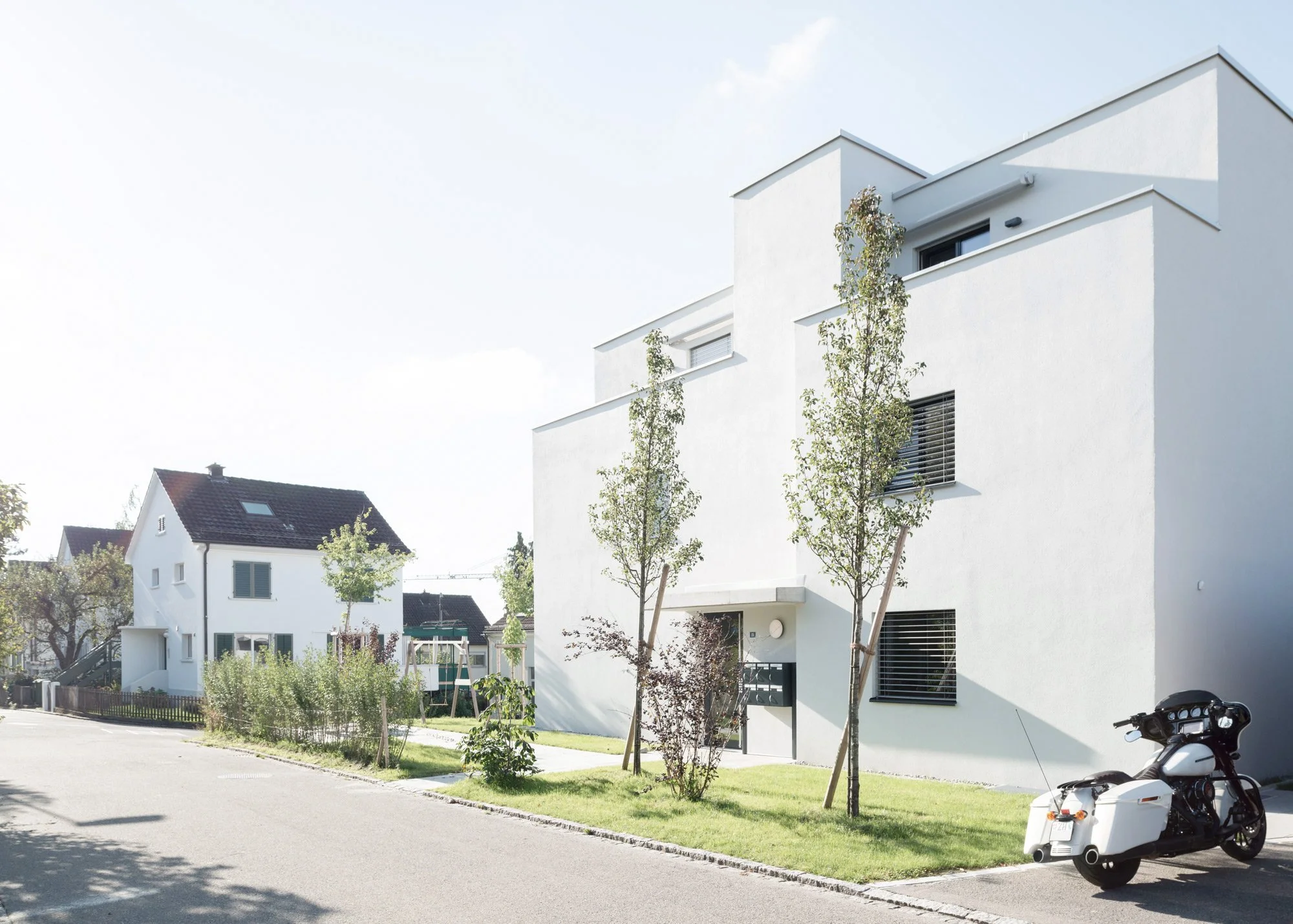 Moderne weisse Mehrfamilienhäuser mit mehreren Etagen und grossen Fenstern, vor einem paar jungen Bäumen.