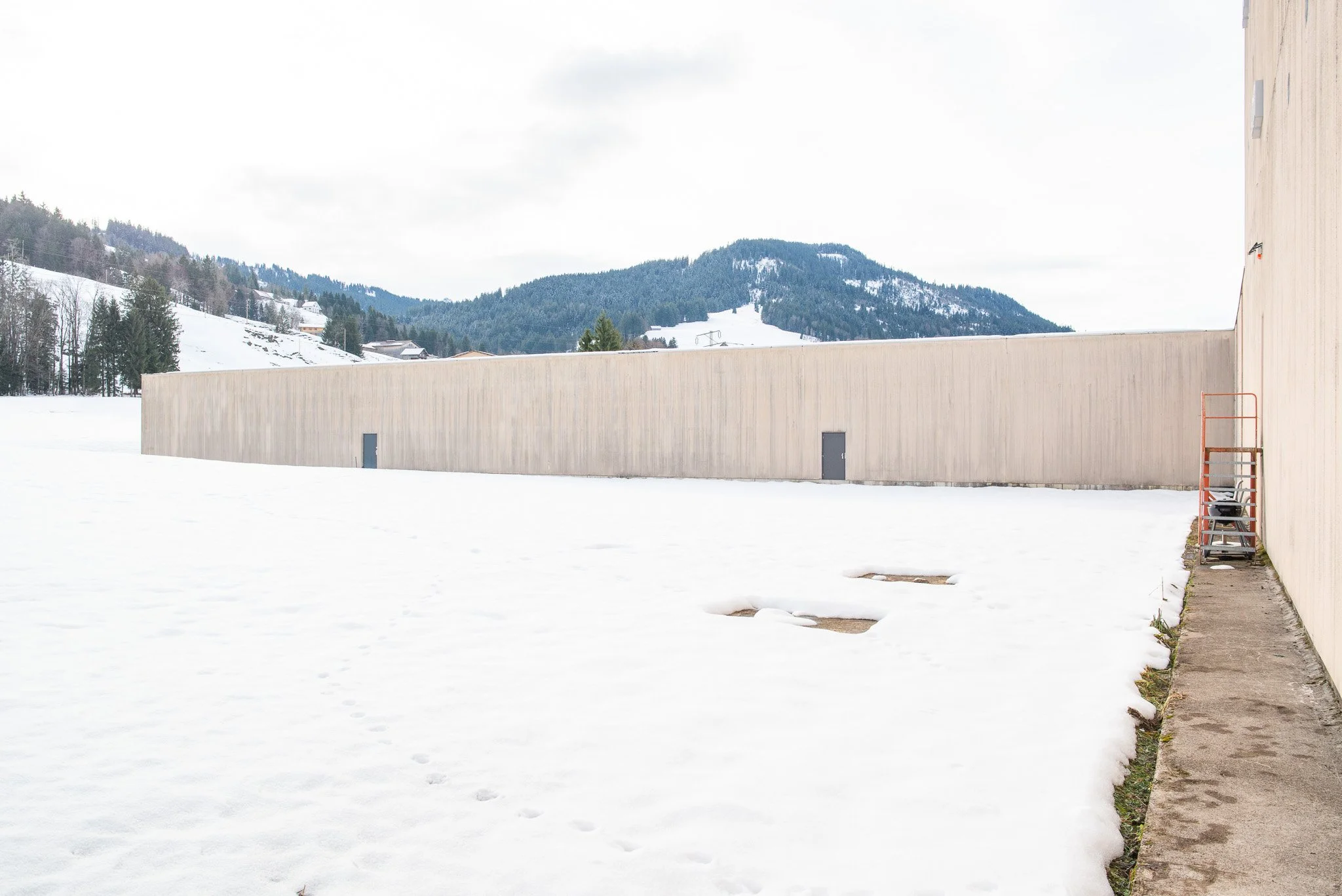 Ein Winterbild zeigt eine weisse, schneebedeckte Ebene mit einer hohen, grauen Mauer links und einem Pfad rechts. Im Hintergrund befinden sich bewaldete Hügel und Berge, unter einem bewölkten Himmel.