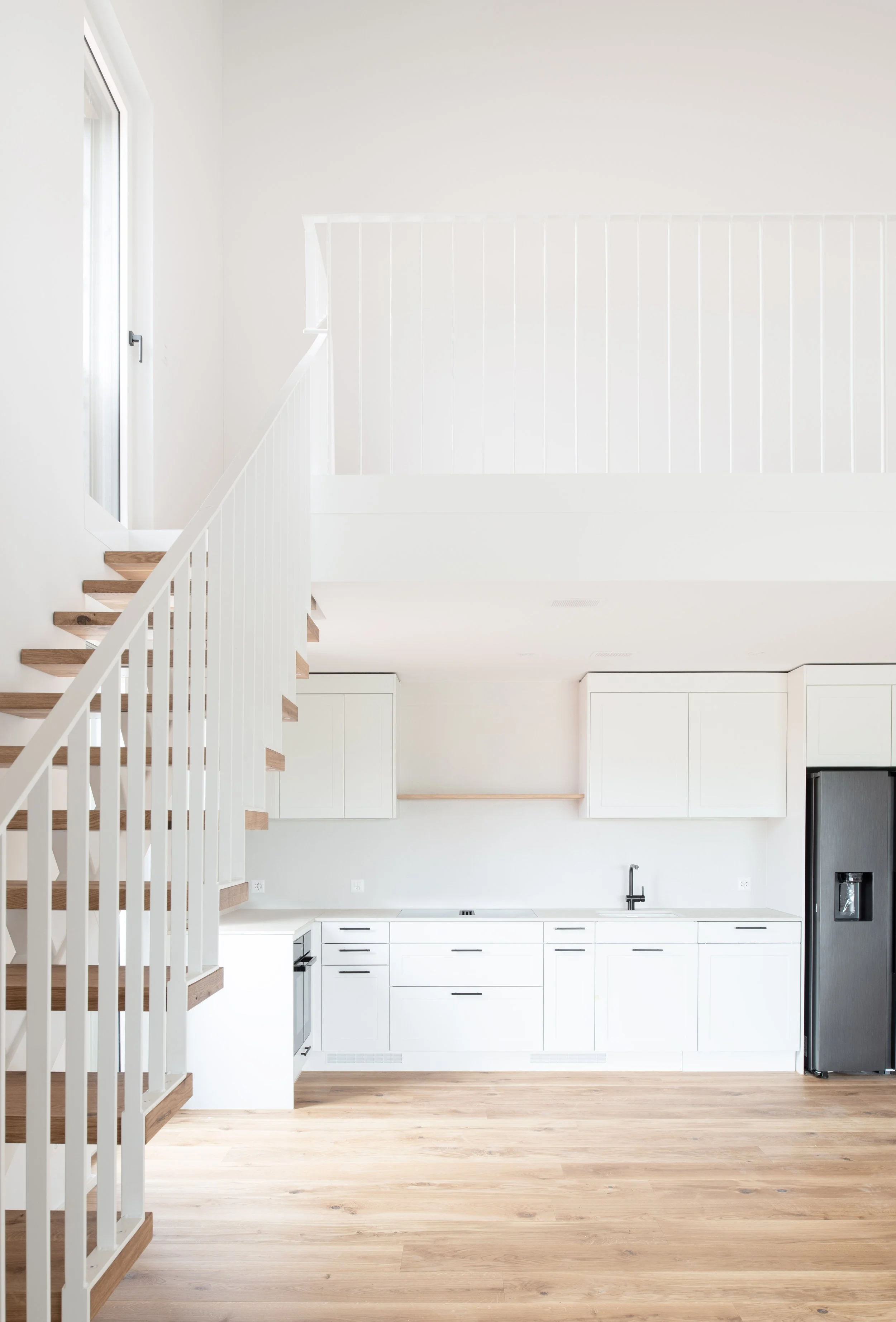Helle, moderne Küche mit weissen Schränken, schwarzer Wasserhahn, Edelstahl-Kühlschrank, Holzfussboden, Hängeschränken, grosse Fenster mit natürlichem Licht, offene Gestaltung, minimalistische Einrichtung.